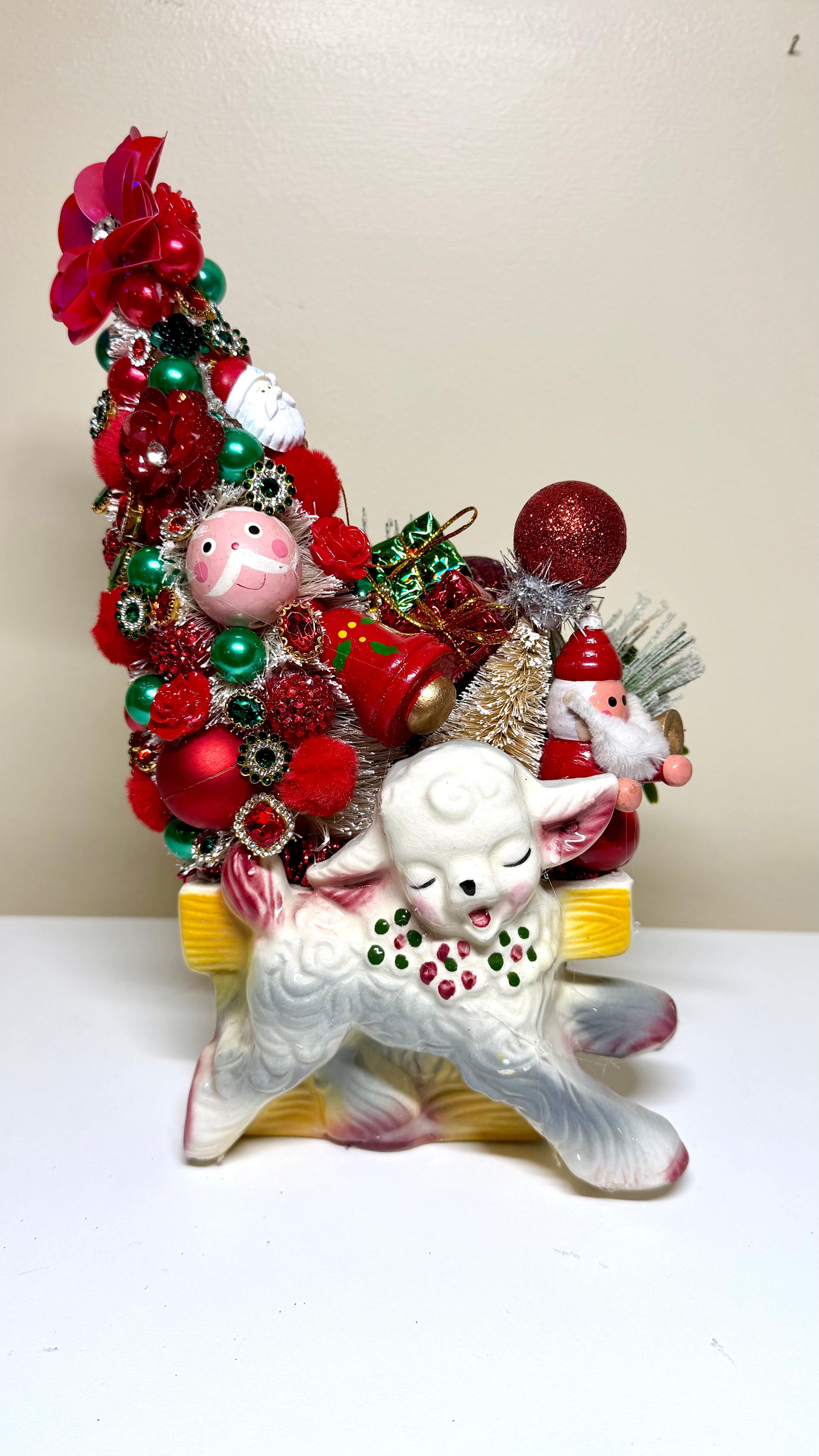 A vintage Christmas ornament display featuring a white ceramic lamb figurine with green and red decorative details, nestled in a large, festive Christmas tree made of red and green ornaments, baubles, and a small Santa Claus figure, set against a plain beige background.