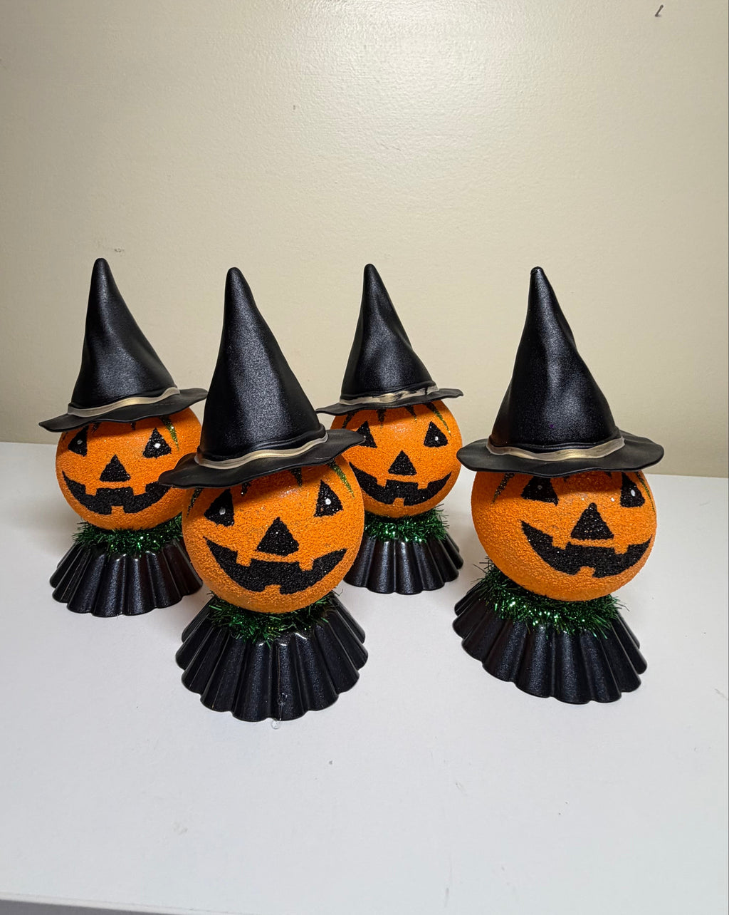 Four Halloween jack-o'-lantern decorations with black witch hats and black pleated skirts, arranged on a white table against a beige wall.