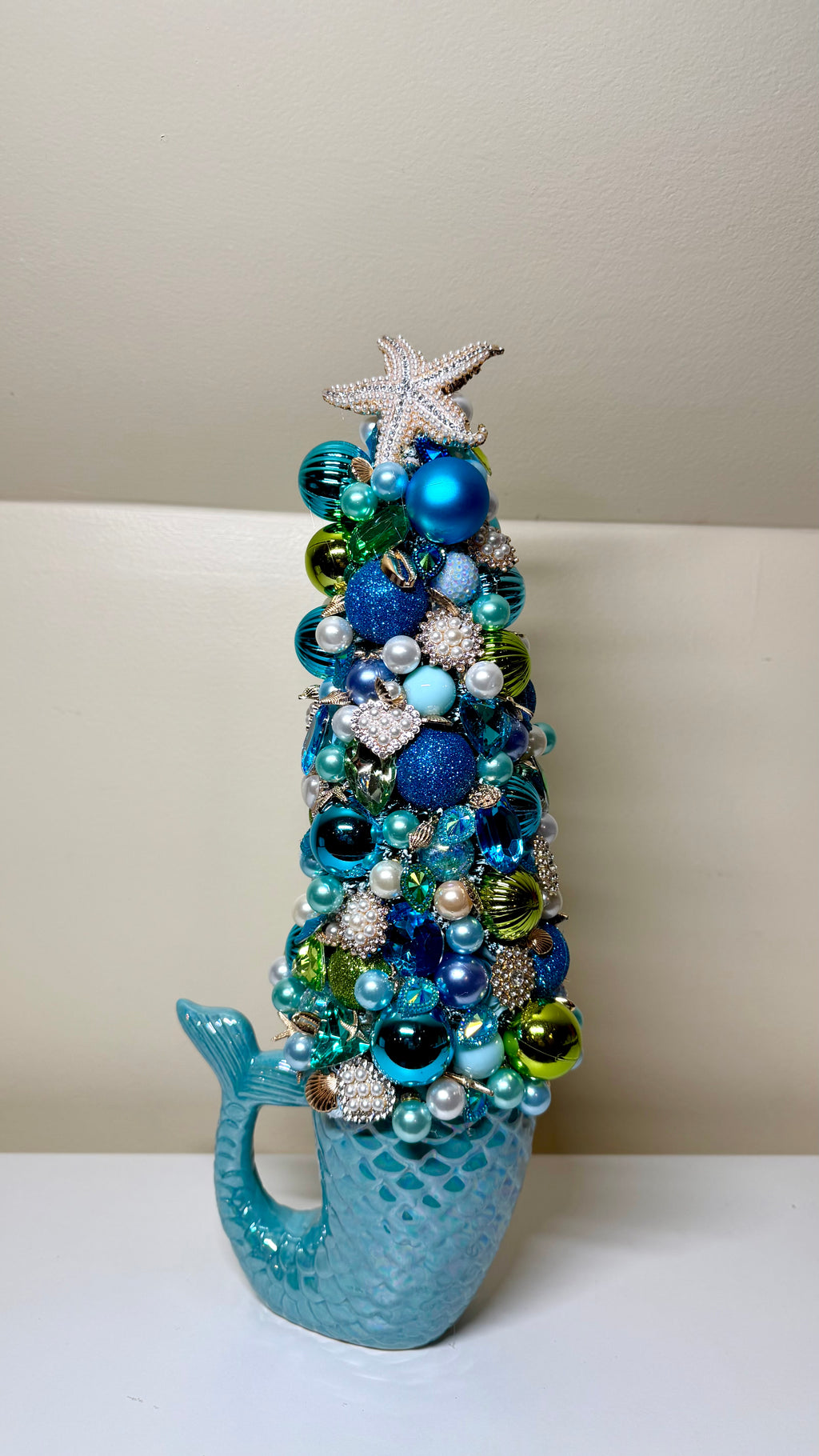 A decorative mermaid-tail-shaped tree ornament filled with blue, teal, green, and silver holiday ornaments and beads, topped with a white starfish, set against a neutral beige background.