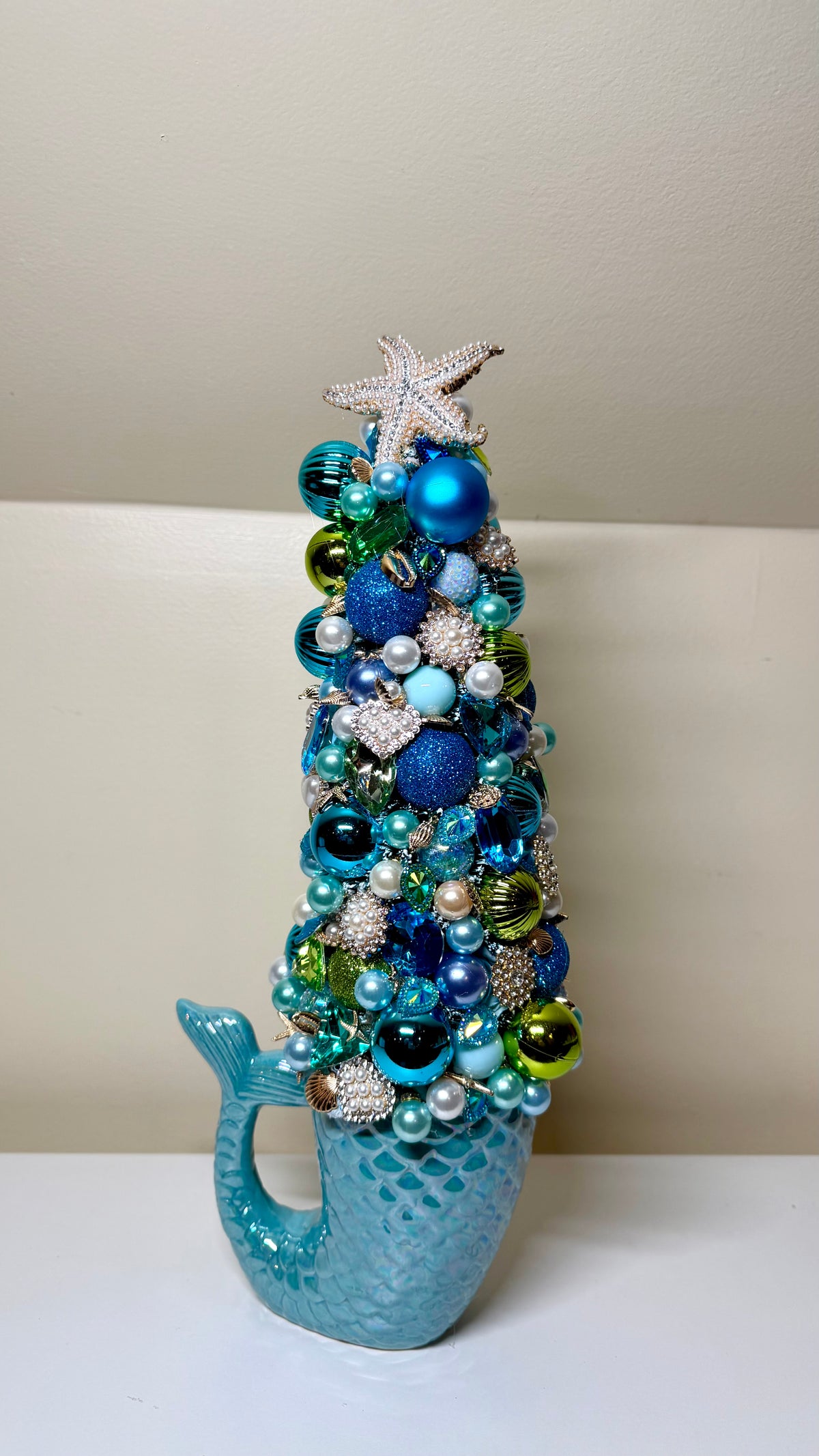 A decorative mermaid-tail-shaped tree ornament filled with blue, teal, green, and silver holiday ornaments and beads, topped with a white starfish, set against a neutral beige background.