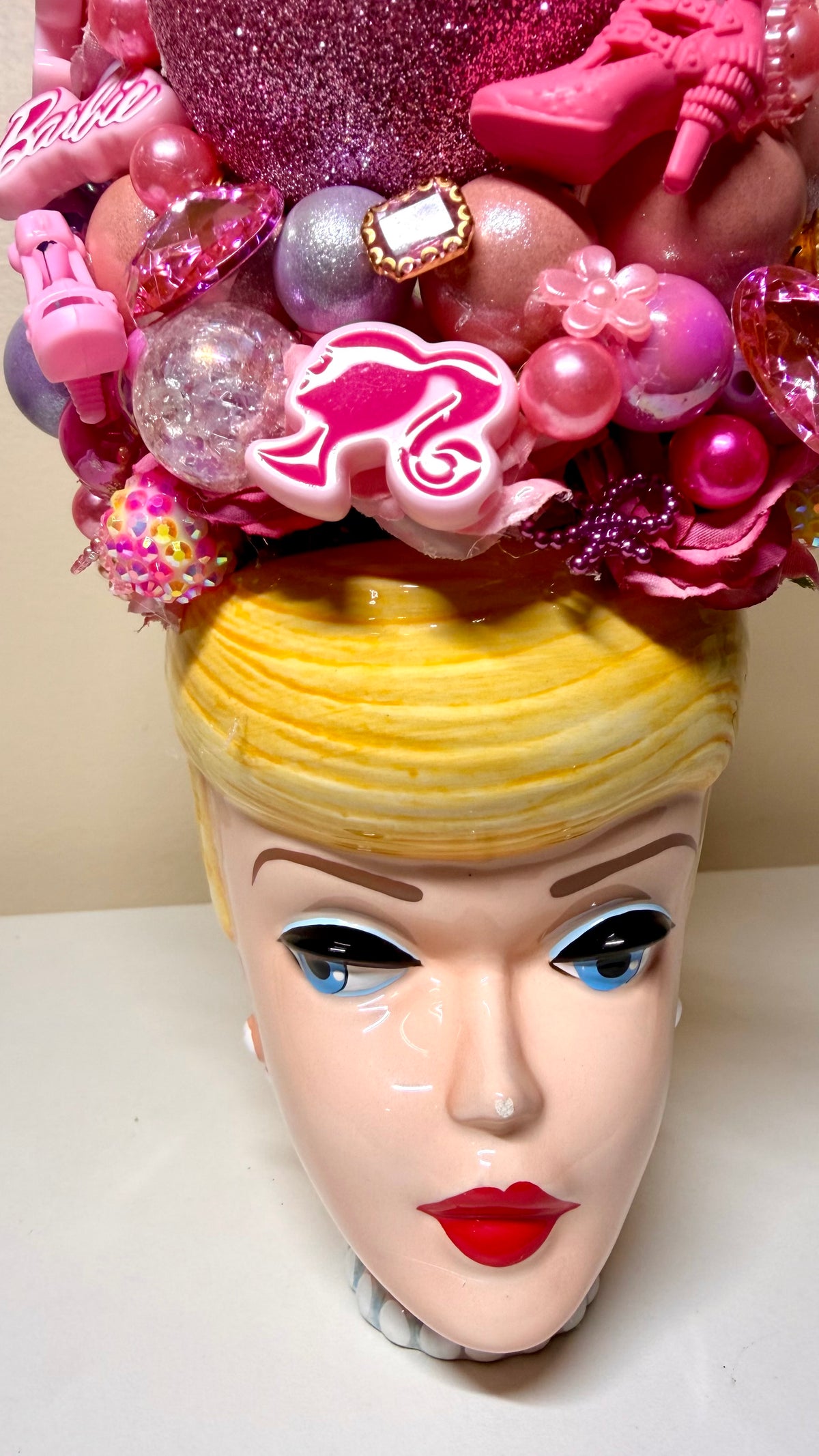A close-up of a decorative Barbie-themed headpiece or hat displayed on a doll-like head. The headpiece is heavily embellished with pink and metallic ornaments, glitter, faux pearls, and pink high-heel charms, while the doll head has bright yellow hair and blue eyes.