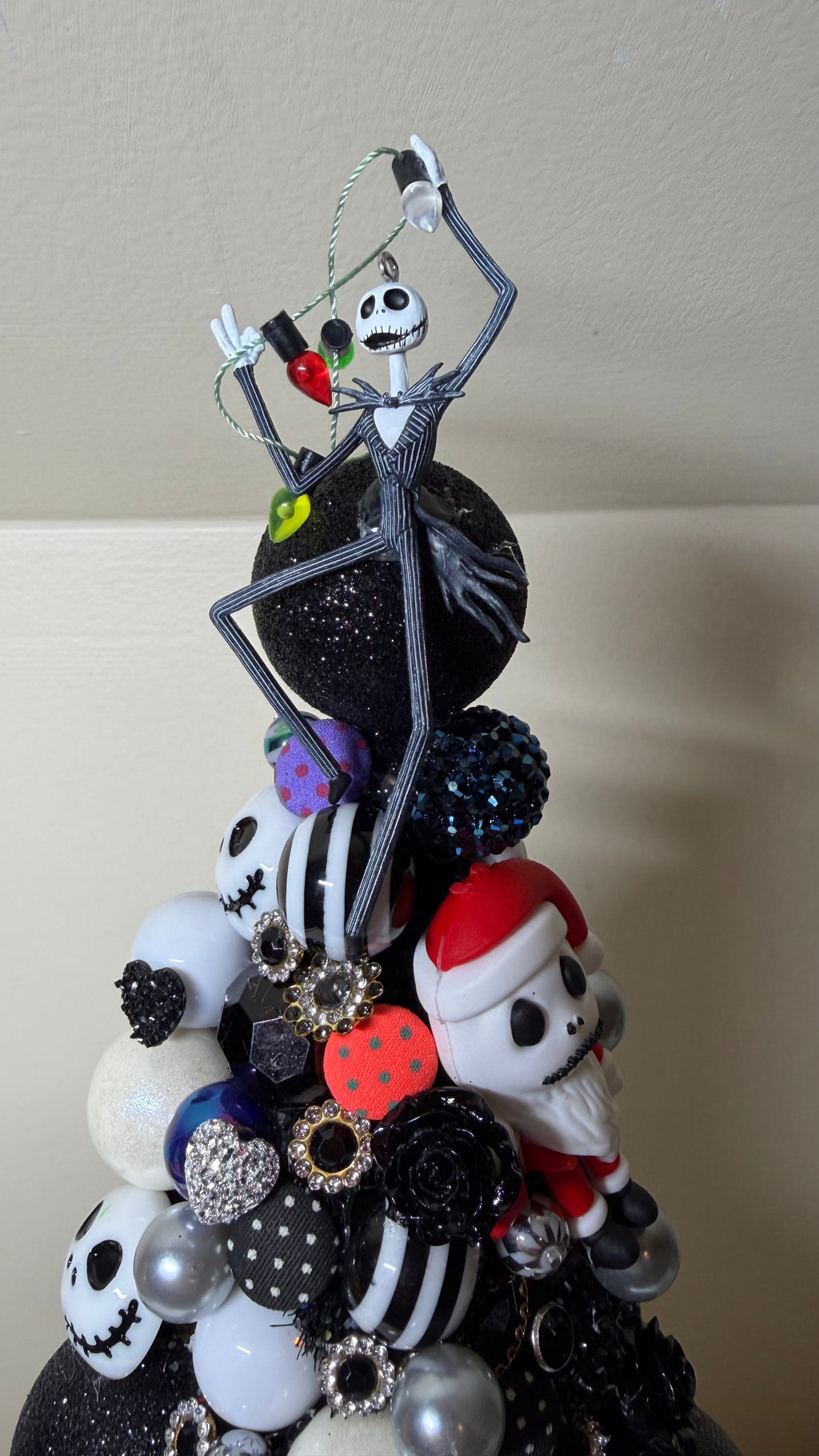 A decorative Christmas tree ornament featuring characters from The Nightmare Before Christmas, including Jack Skellington, Sally, and Santa, arranged on a black glittery base with black, white, and red ornaments.