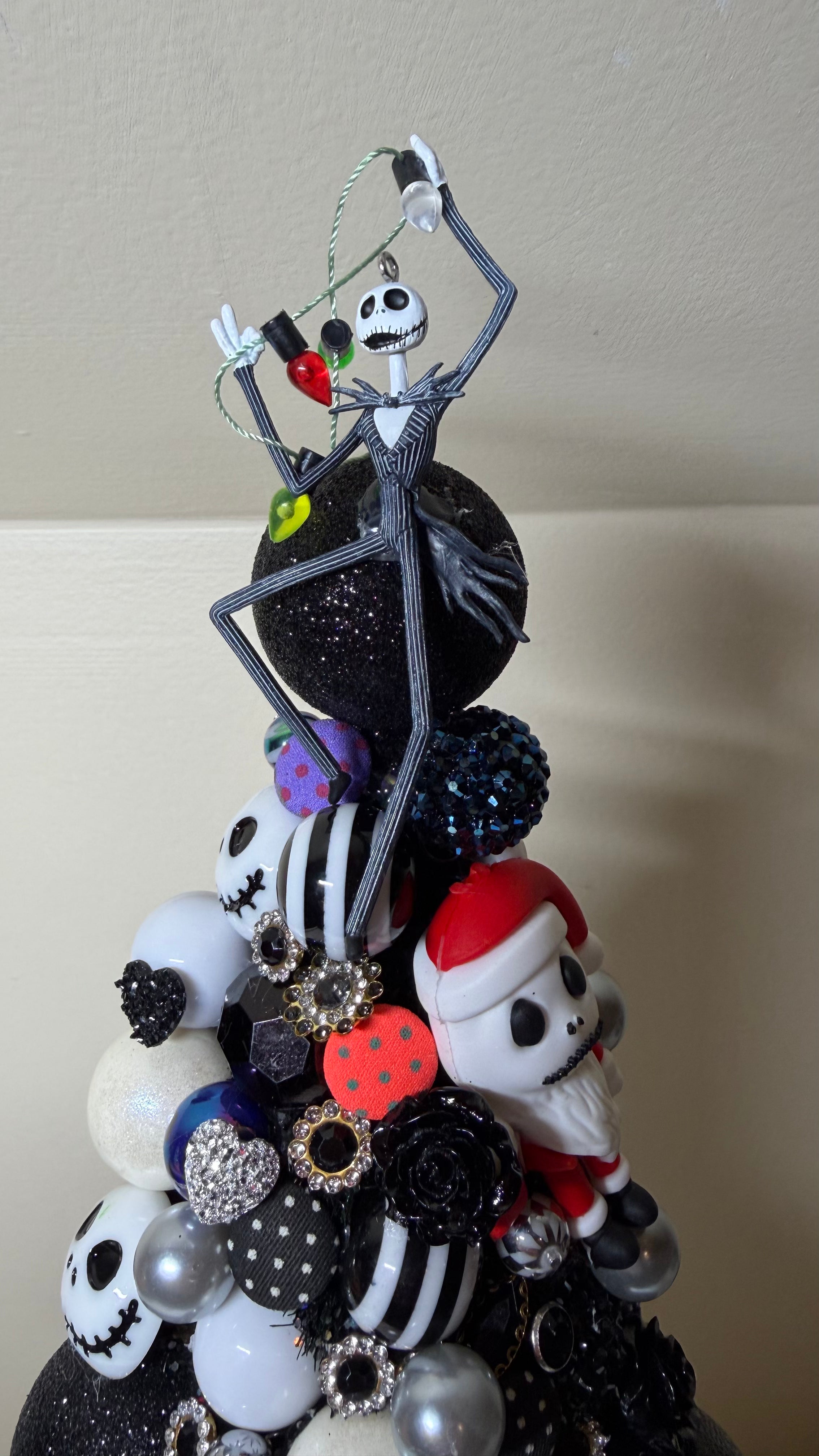 A decorative Christmas tree ornament featuring characters from The Nightmare Before Christmas, including Jack Skellington, Sally, and Santa, arranged on a black glittery base with black, white, and red ornaments.