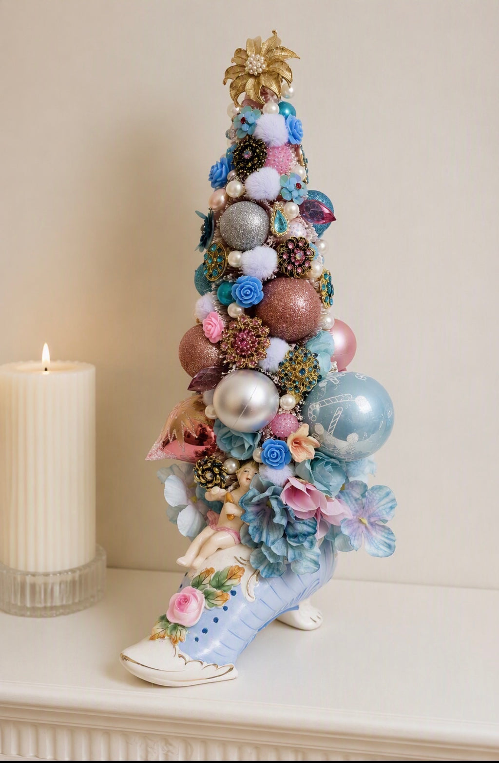 A decorative miniature Christmas tree shaped like a high-heeled shoe, decorated with pastel blue and pink flowers, silver and pink ornaments, and a gold star topper, displayed on a white mantel next to a lit white candle.