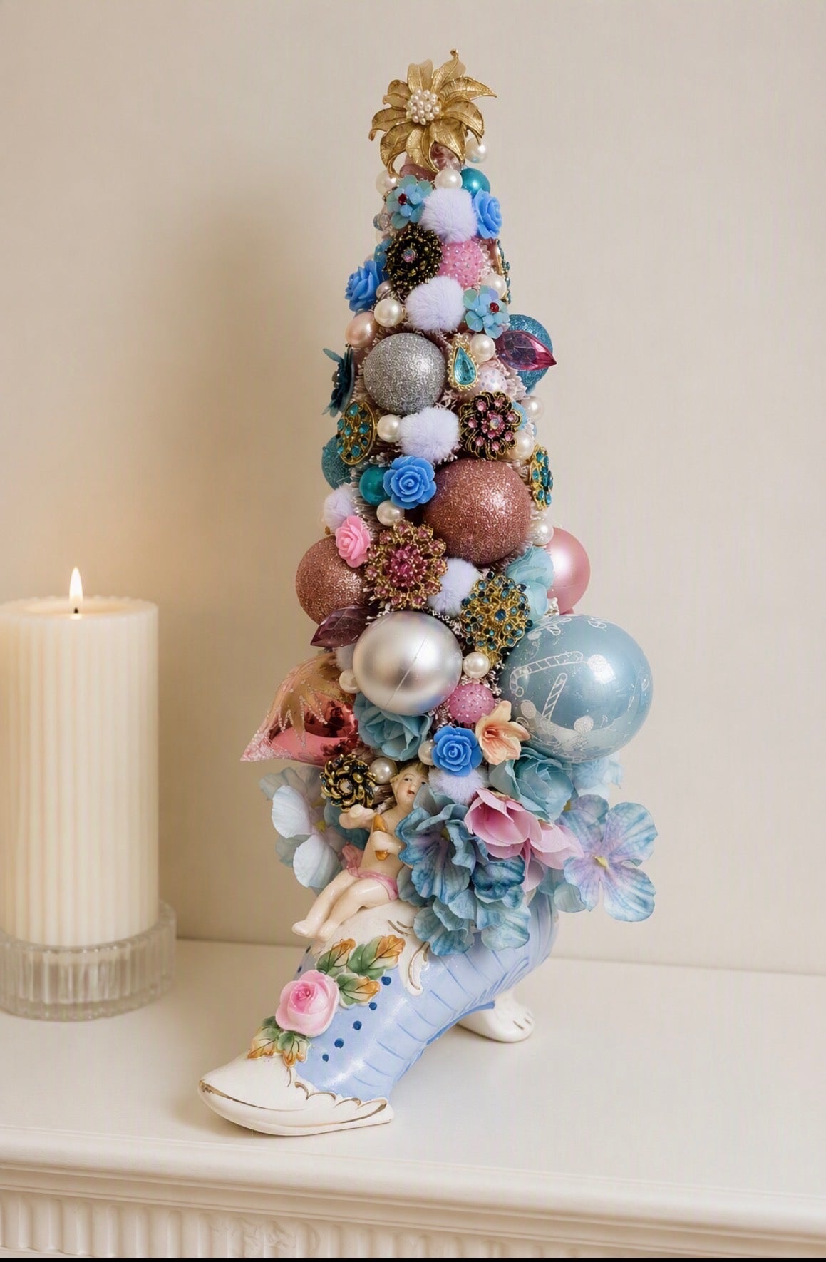 A decorative miniature Christmas tree shaped like a high-heeled shoe, decorated with pastel blue and pink flowers, silver and pink ornaments, and a gold star topper, displayed on a white mantel next to a lit white candle.