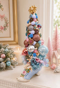 A decorative Christmas tree shaped like a light blue high-heeled shoe, decorated with pastel pom-poms, fabric flowers, glittery ornaments, and a gold star topper, displayed on a mantel in a softly lit room.