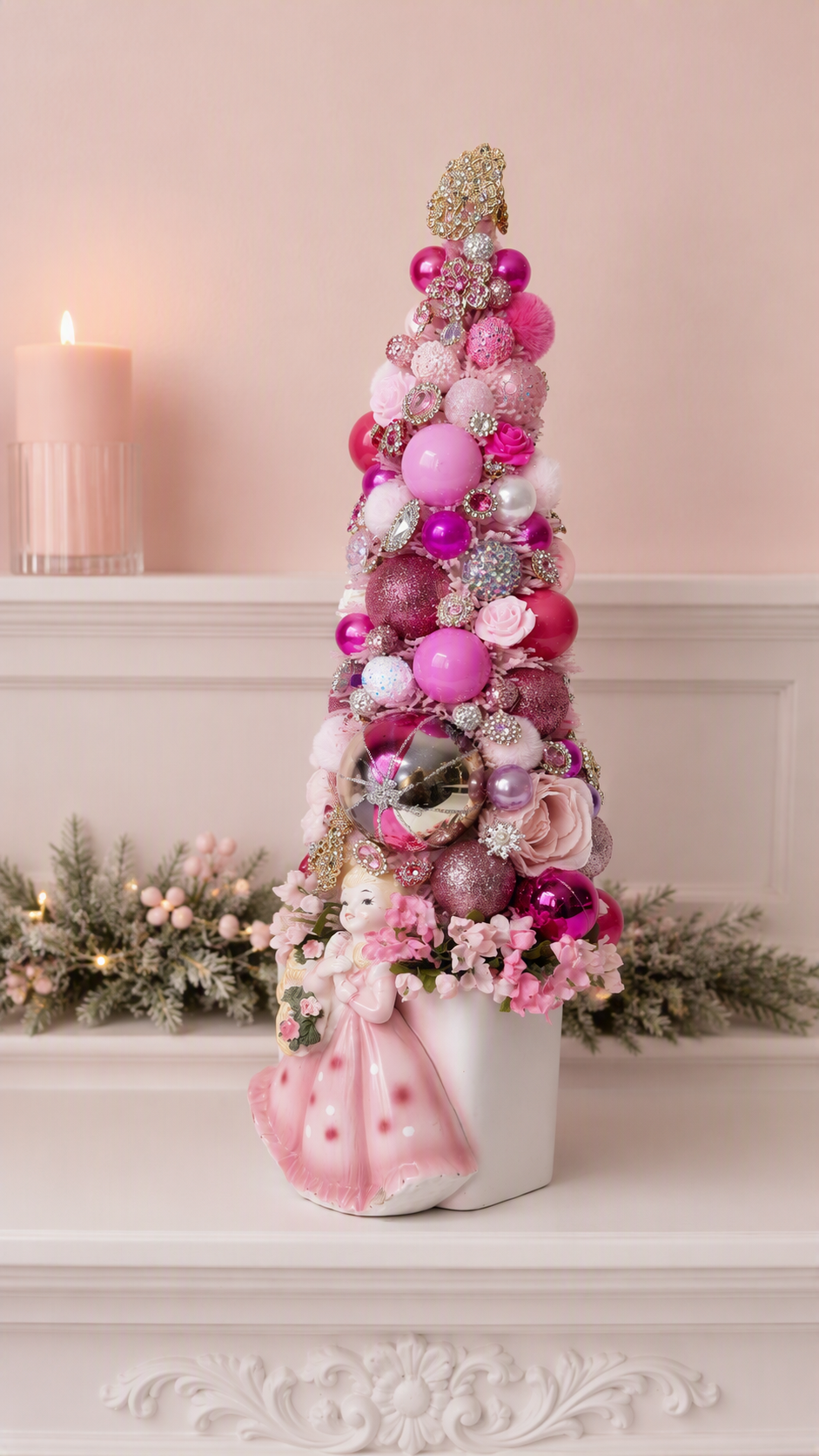 A decorative miniature Christmas tree arrangement in a white pot, decorated with pink and silver ornaments, glittery pink ribbons, and a pink bow, displayed on a white mantel against a soft pink wall.