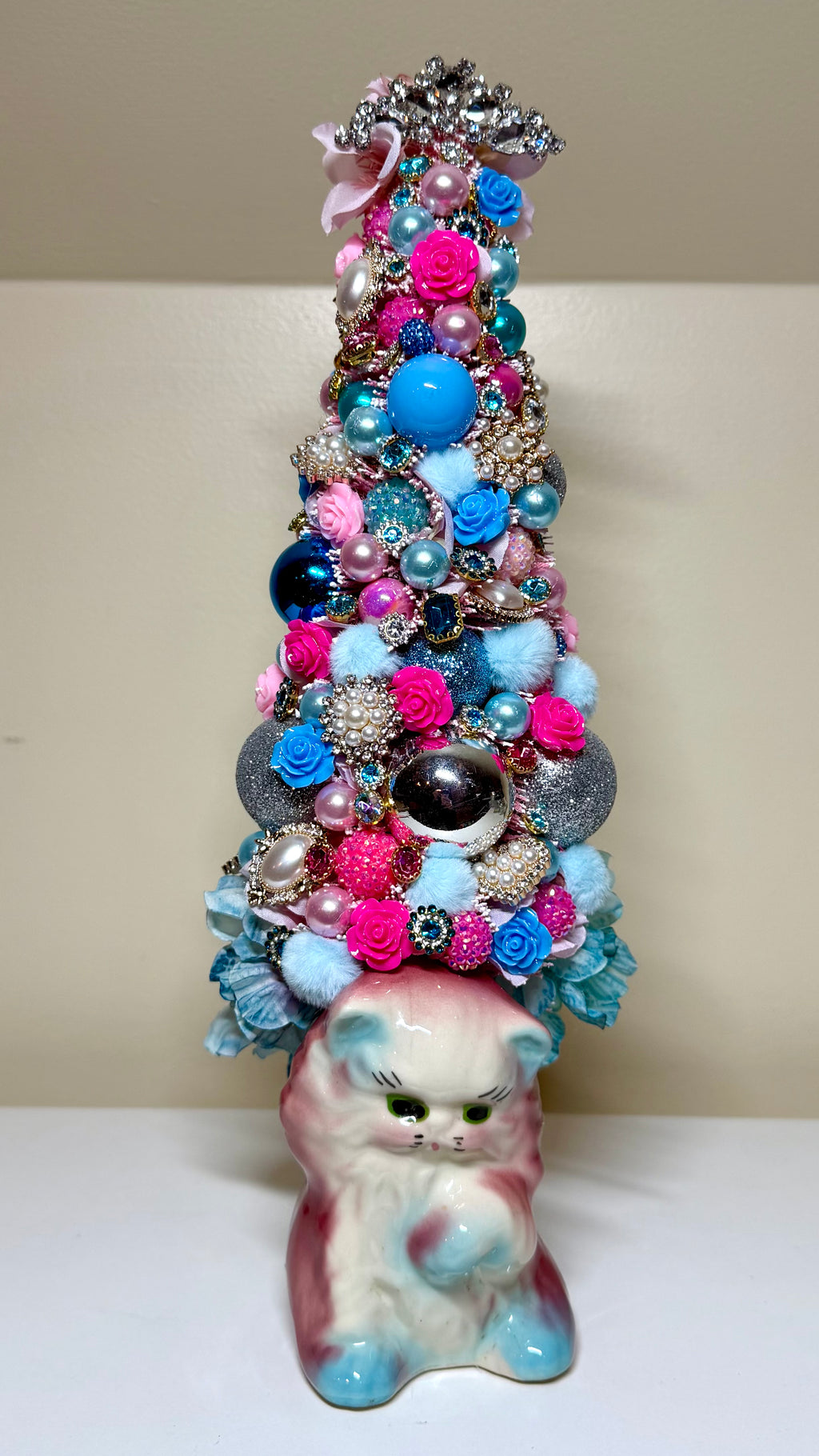 A decorative Christmas tree made from layered brooches and jewelry ornaments, topped with a silver star and pink bow, standing on a pink and blue ceramic kitten figurine against a beige background.