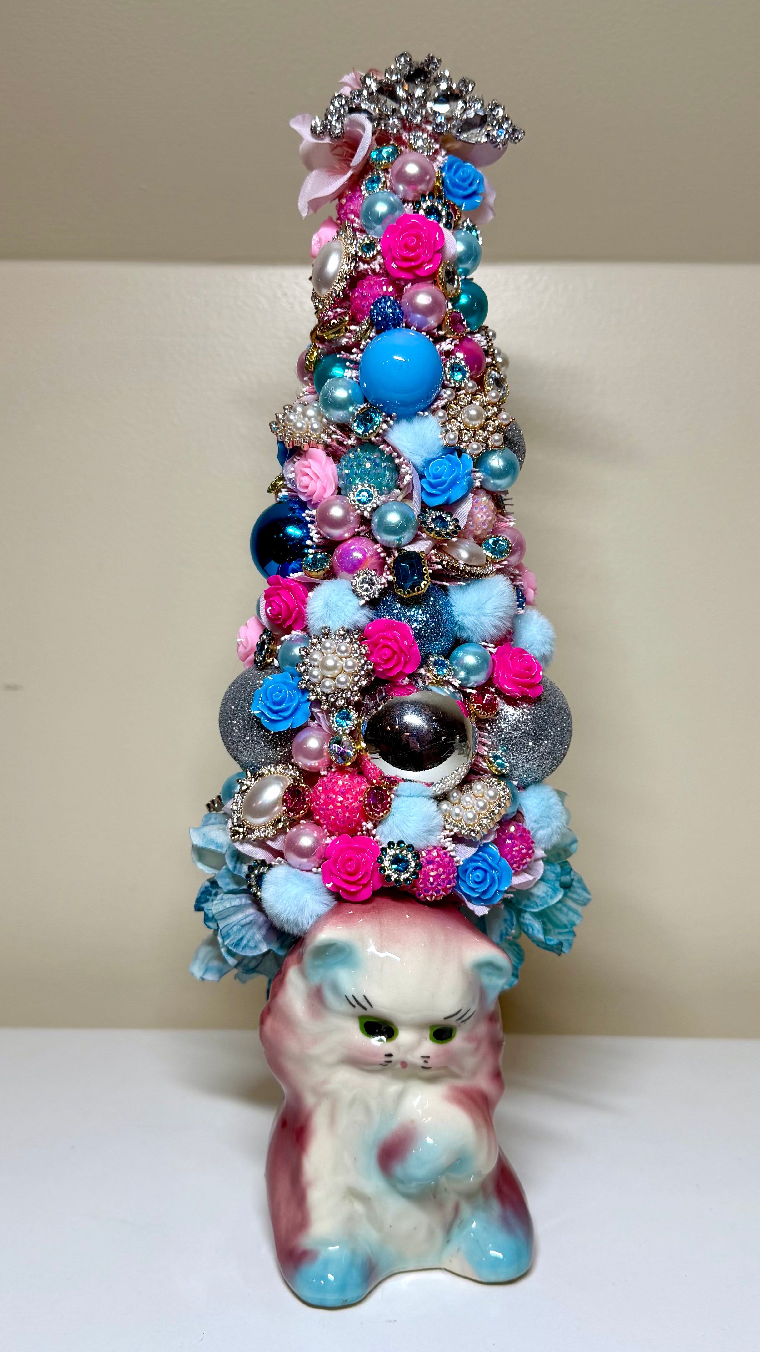 A decorative Christmas tree made from layered brooches and jewelry ornaments, topped with a silver star and pink bow, standing on a pink and blue ceramic kitten figurine against a beige background.