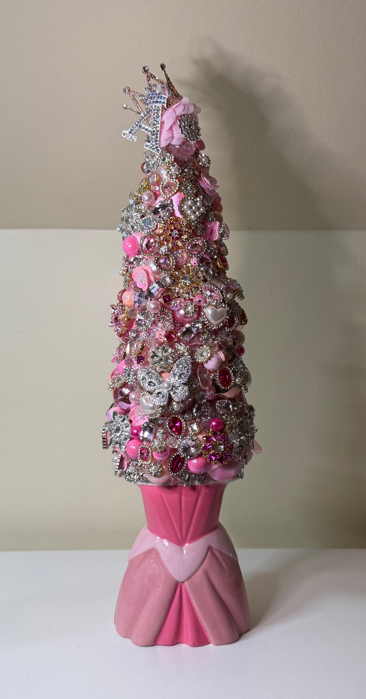 A decorative Christmas tree crafted from pink and silver rhinestones, sequins, and beads, mounted on a pink satin skirt-shaped base against a beige wall.
