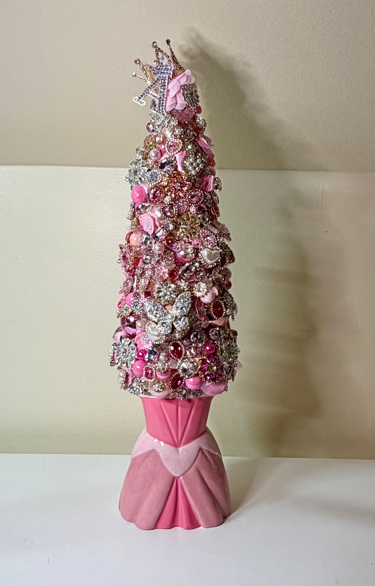 A pink and silver Christmas tree ornament tree with a crown topper, decorated with pink and silver ornaments, displayed on a white surface against a beige wall.