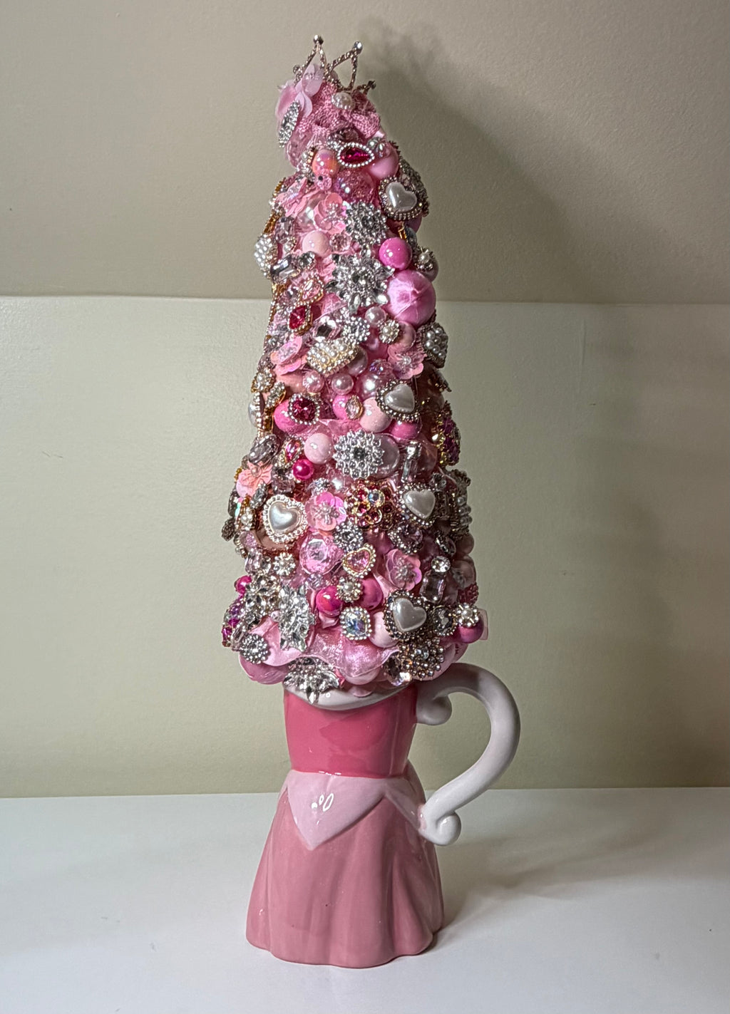 A pink ceramic mug shaped like a princess skirt with a handle, topped with a decorative Christmas tree made of pink and silver ornaments, beads, and sequins, set against a plain beige wall and white surface.