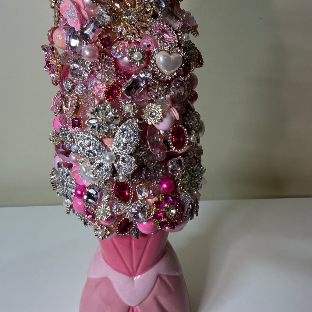 A pink and silver decorative bust or vase-like object covered densely with rhinestones, brooches, pearls, and jeweled embellishments, photographed against a plain indoor background.