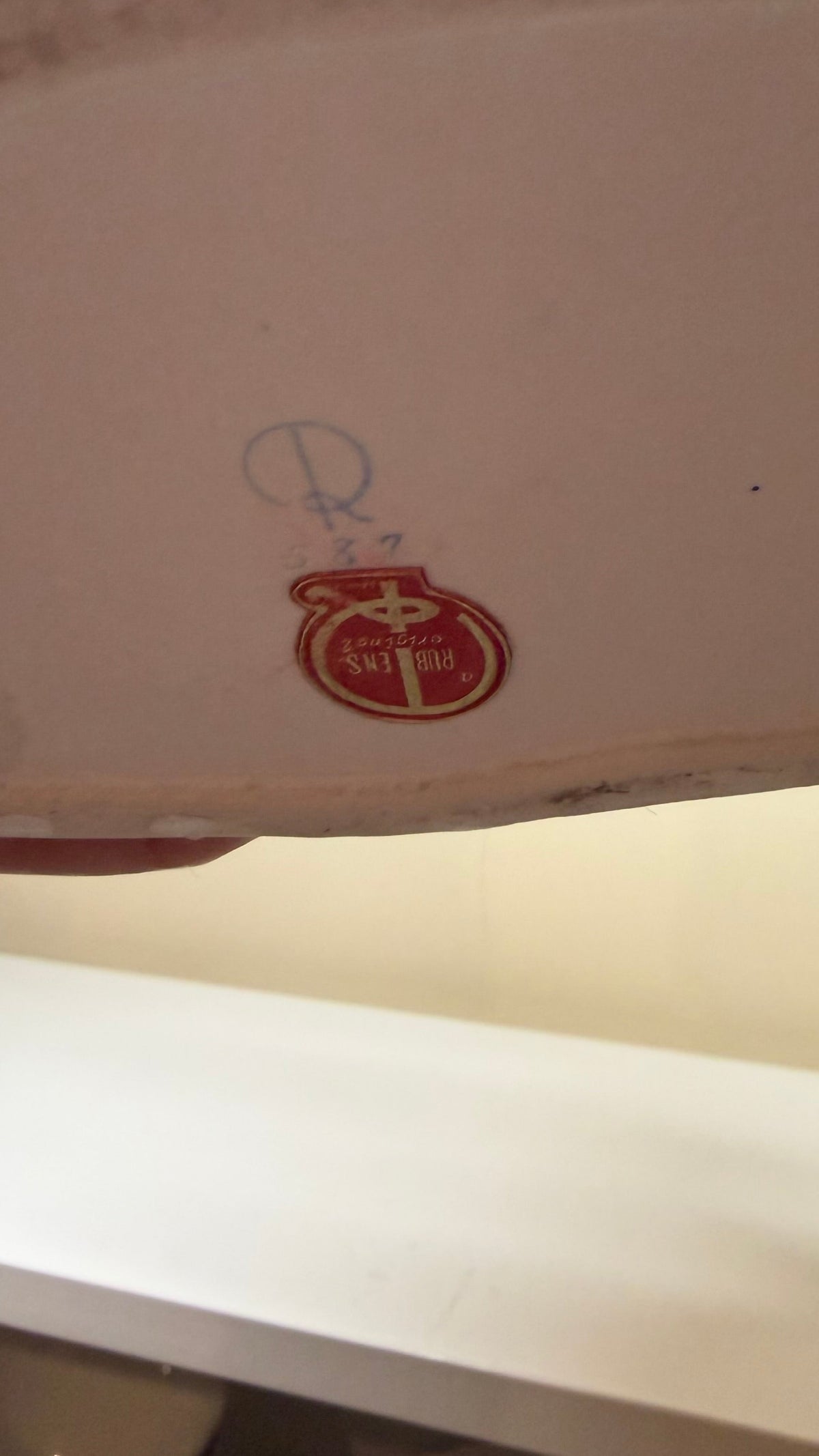 A close-up of the bottom edge of a white ceramic plate or dish, showing a small red and gold circular logo and a blue registration mark. The plate is held in front of a white shelf in a kitchen setting.