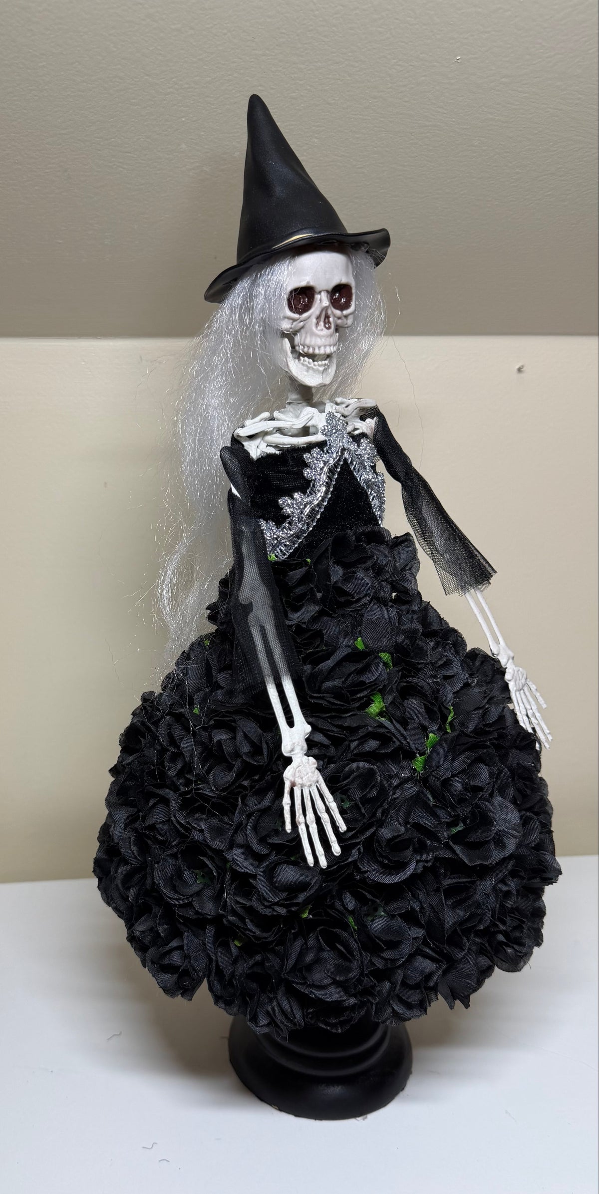 A Halloween skeleton witch figurine wearing a black pointed hat and a black rose-covered dress, displayed on a white surface against a beige wall.