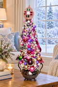 A decorative tabletop Christmas tree filled with colorful ornaments in pink, purple, silver, white, and green, displayed on a wooden table in a cozy living room near a window showing a snowy outdoor view.
