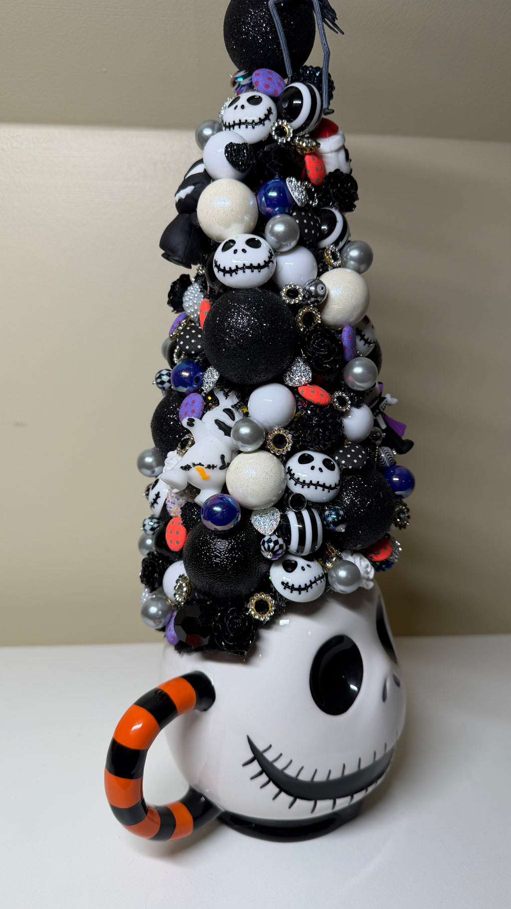 A decorative Halloween tree made from a white mug with a cartoon face, covered in black, white, and silver ornaments, beads, and small skull figures, with an orange-and-black striped handle.