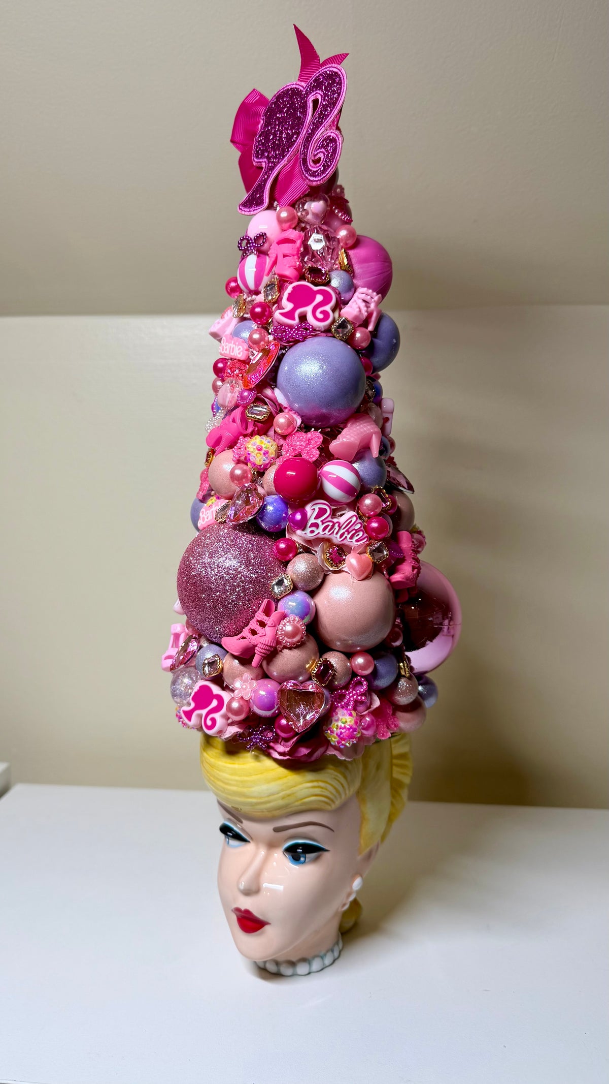 A Barbie-themed decorative headpiece or hat displayed on a stylized blonde doll head. The tall, cone-shaped accessory is covered in pink and purple ornaments, beads, glitter, and small Barbie logo charms, photographed against a plain beige wall and white surface.