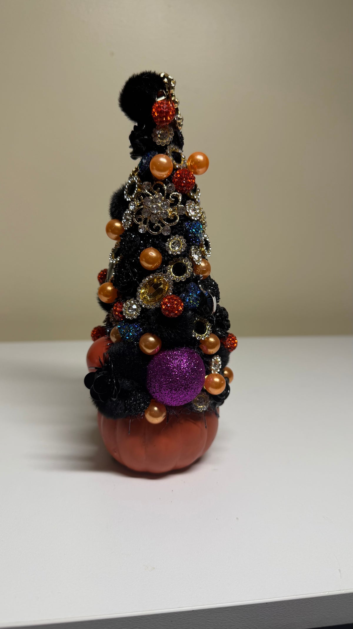 A small orange pumpkin decoration shaped like a festive Christmas tree, decorated with black pom-poms, orange and gold beads, glittery orange and purple ornaments, and a black pom-pom topper, standing on a white surface against a beige background.