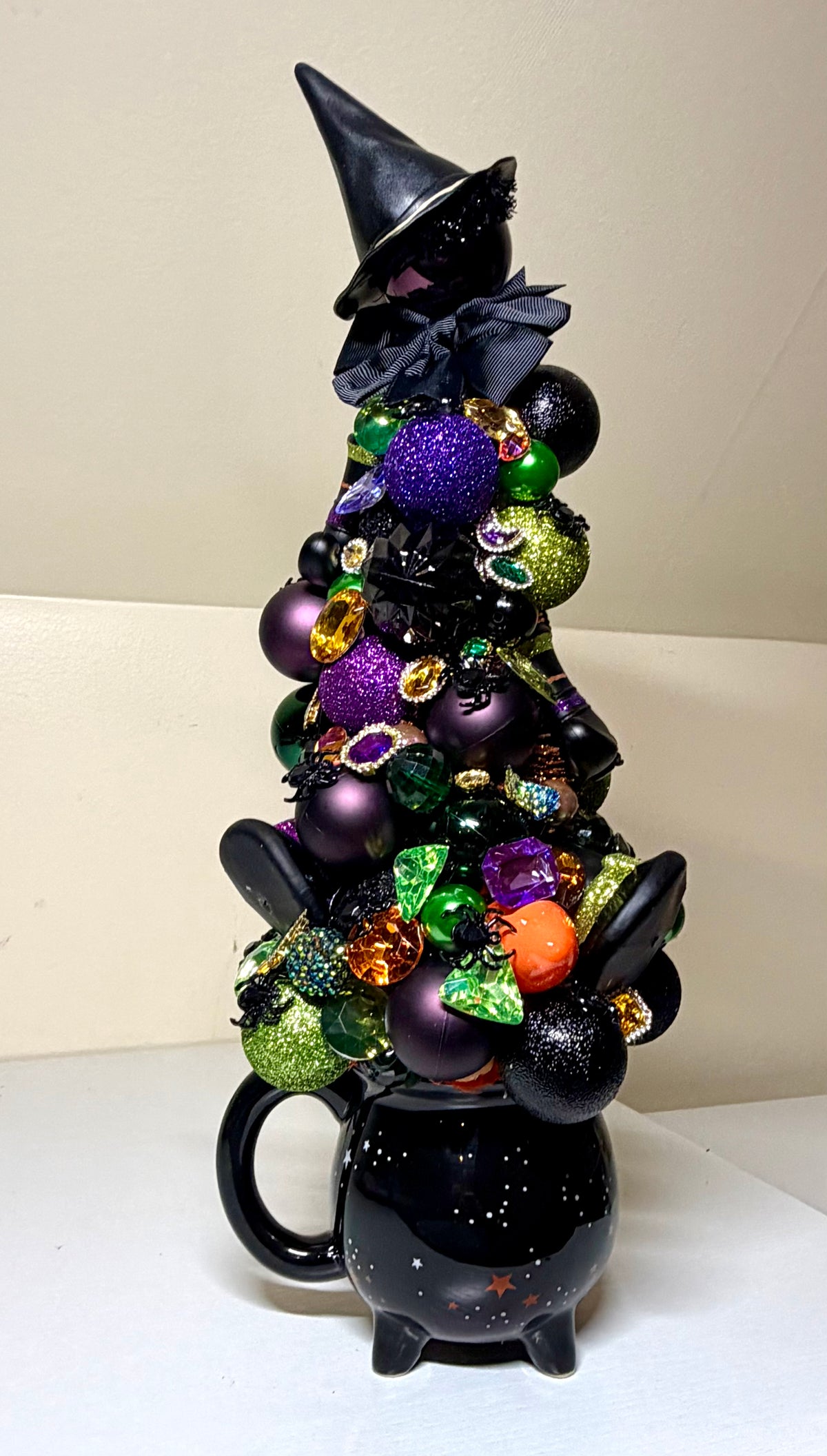 A decorative black witch cauldron figurine topped with a black witch hat and adorned with purple, green, and black glittery ornaments, set against a plain beige background.