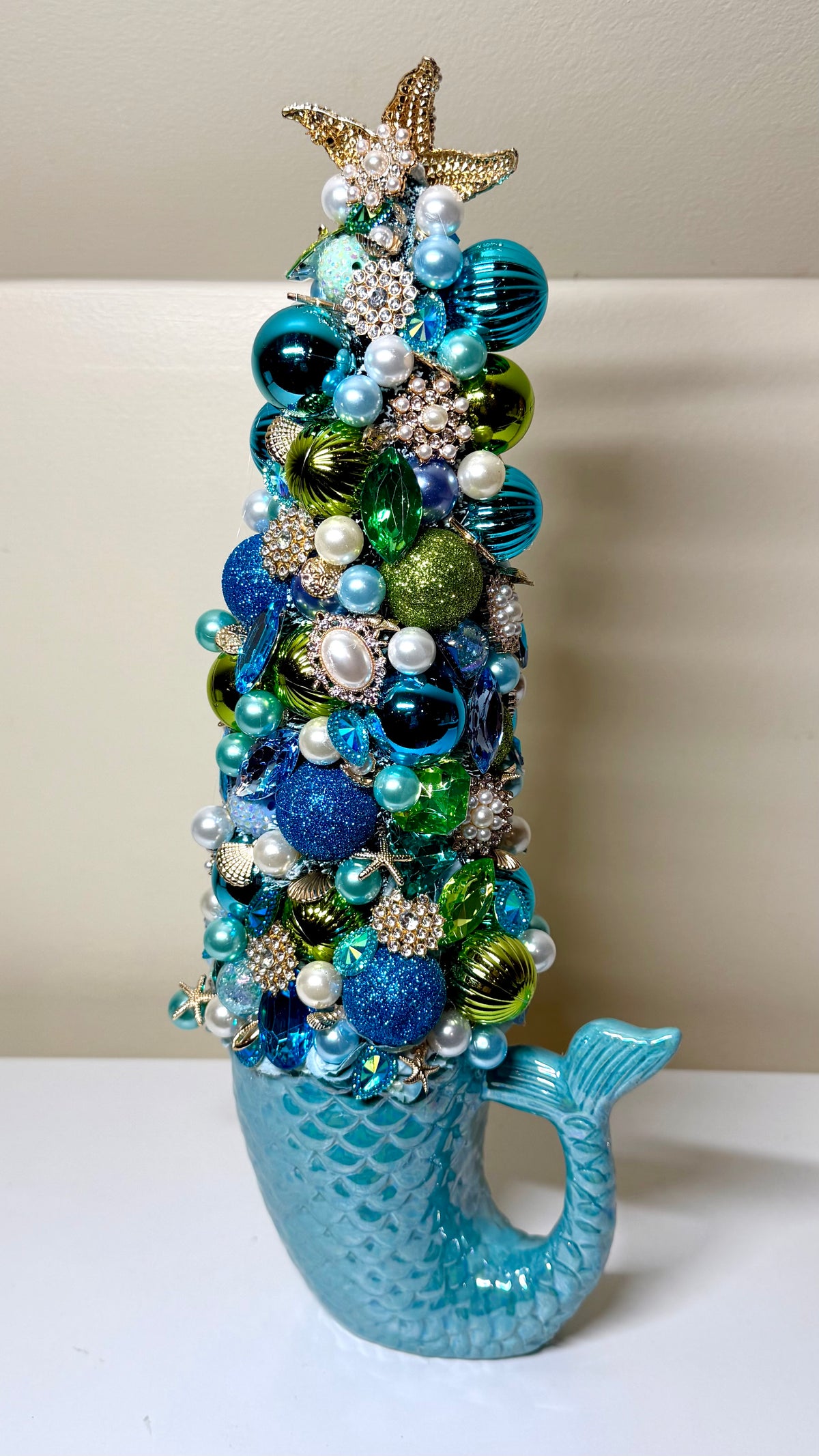 A decorative mermaid-tail-shaped tree ornament covered with blue, teal, green, and white beads, pearls, rhinestones, and starfish toppers, photographed against a plain beige wall and white surface.