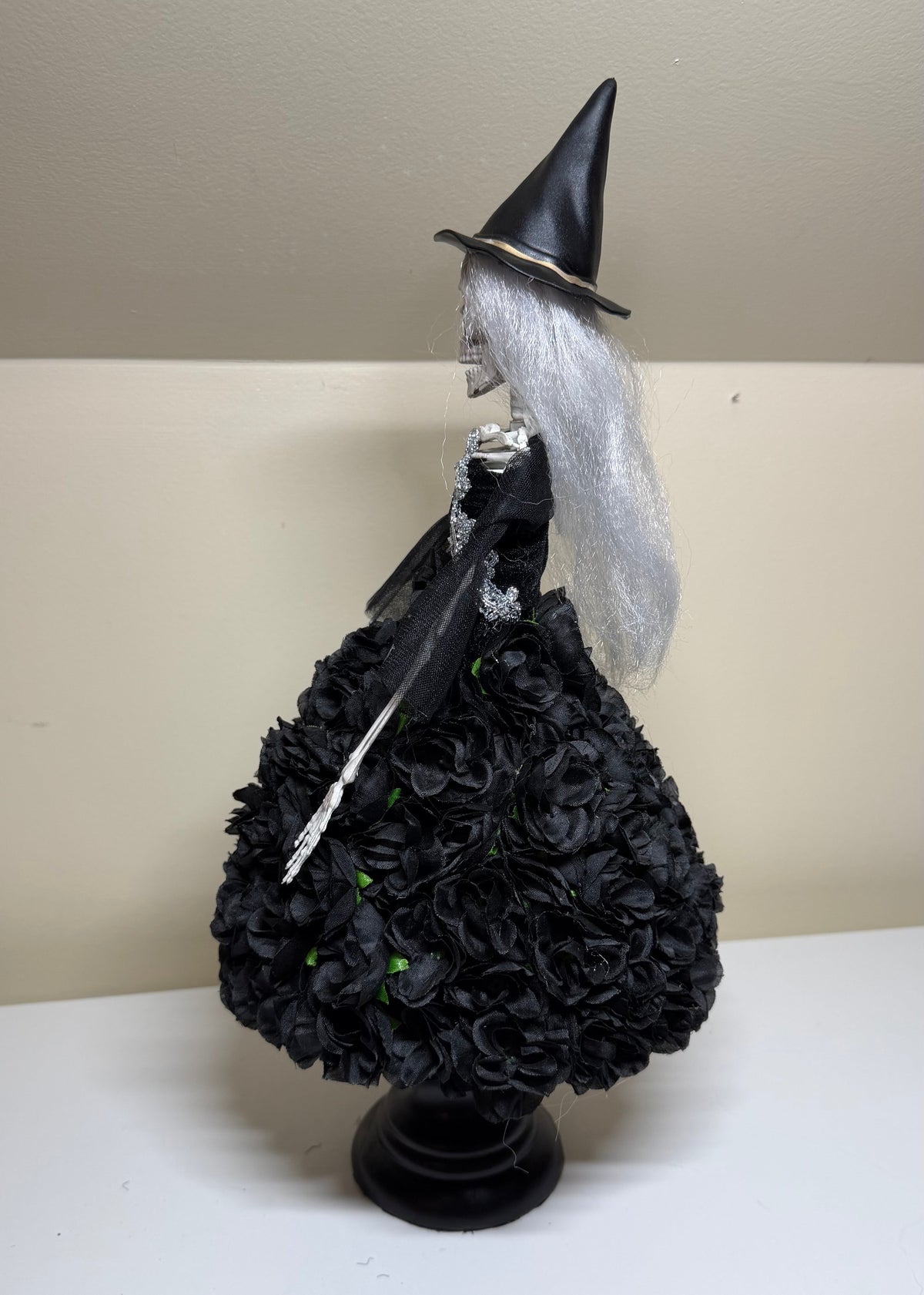 A Halloween witch figurine with a black pointed hat, long white hair, and a black rose-covered dress, standing on a black base against a beige wall and white shelf.