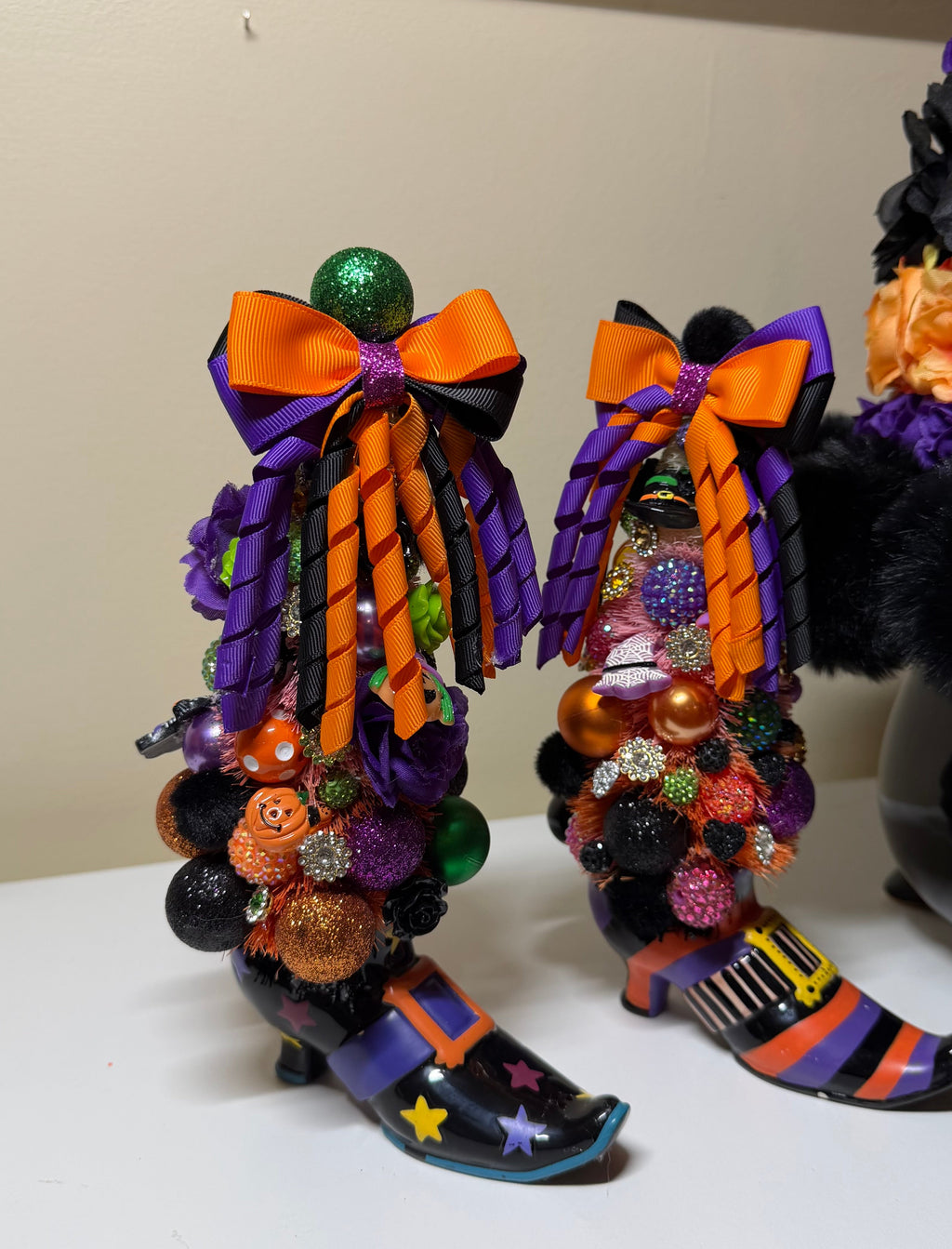 Two decorative witch-style boot ornaments with large orange and purple bows, multicolored pom-poms, beads, and striped orange and purple bases, displayed on a white surface against a beige wall.