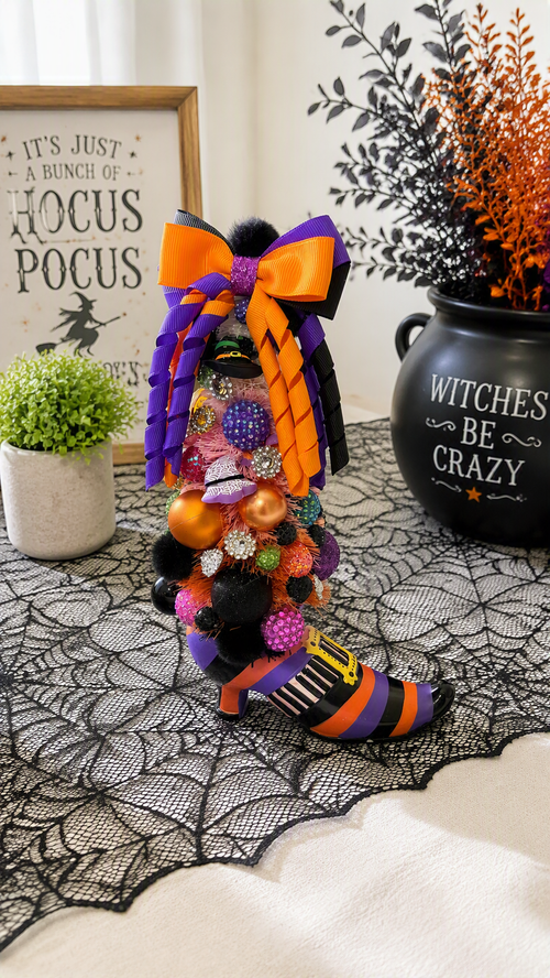 A decorative witch boot ornament filled with colorful beads and ornaments, styled on a black lace runner with Halloween-themed props including a black pot, a small green plant, and a framed sign.