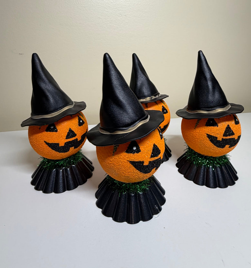 Four orange pumpkin-shaped Halloween decorations with carved jack-o'-lantern faces, black pointed witch hats, and black ruffled skirts, arranged on a white surface against a plain beige wall.