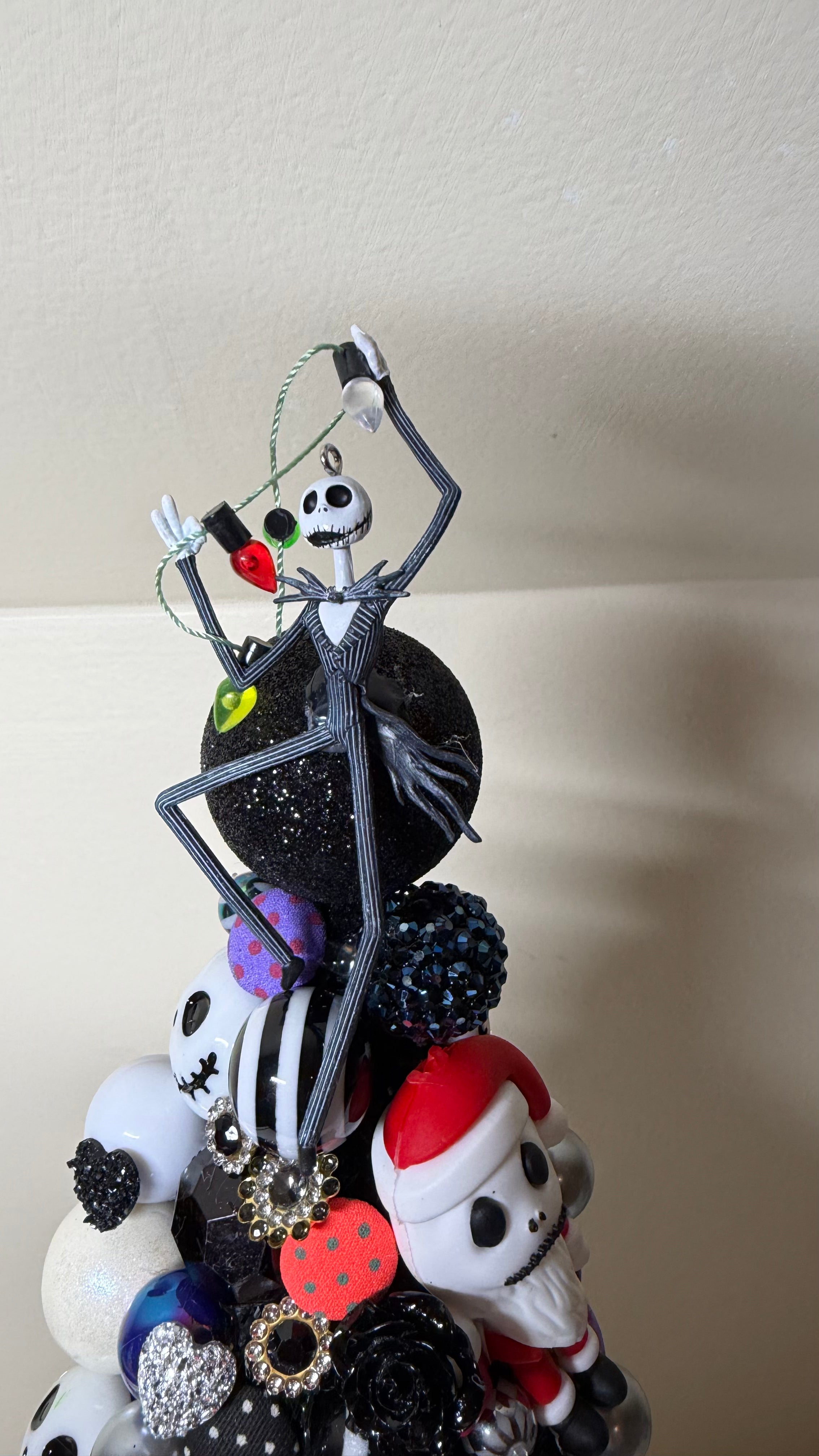 A decorative Christmas tree topper featuring Jack Skellington perched on a black glittery sphere above a cluster of small skull ornaments, black and white polka dots, and red-and-white Santa-style figures, photographed indoors against a plain beige wall.