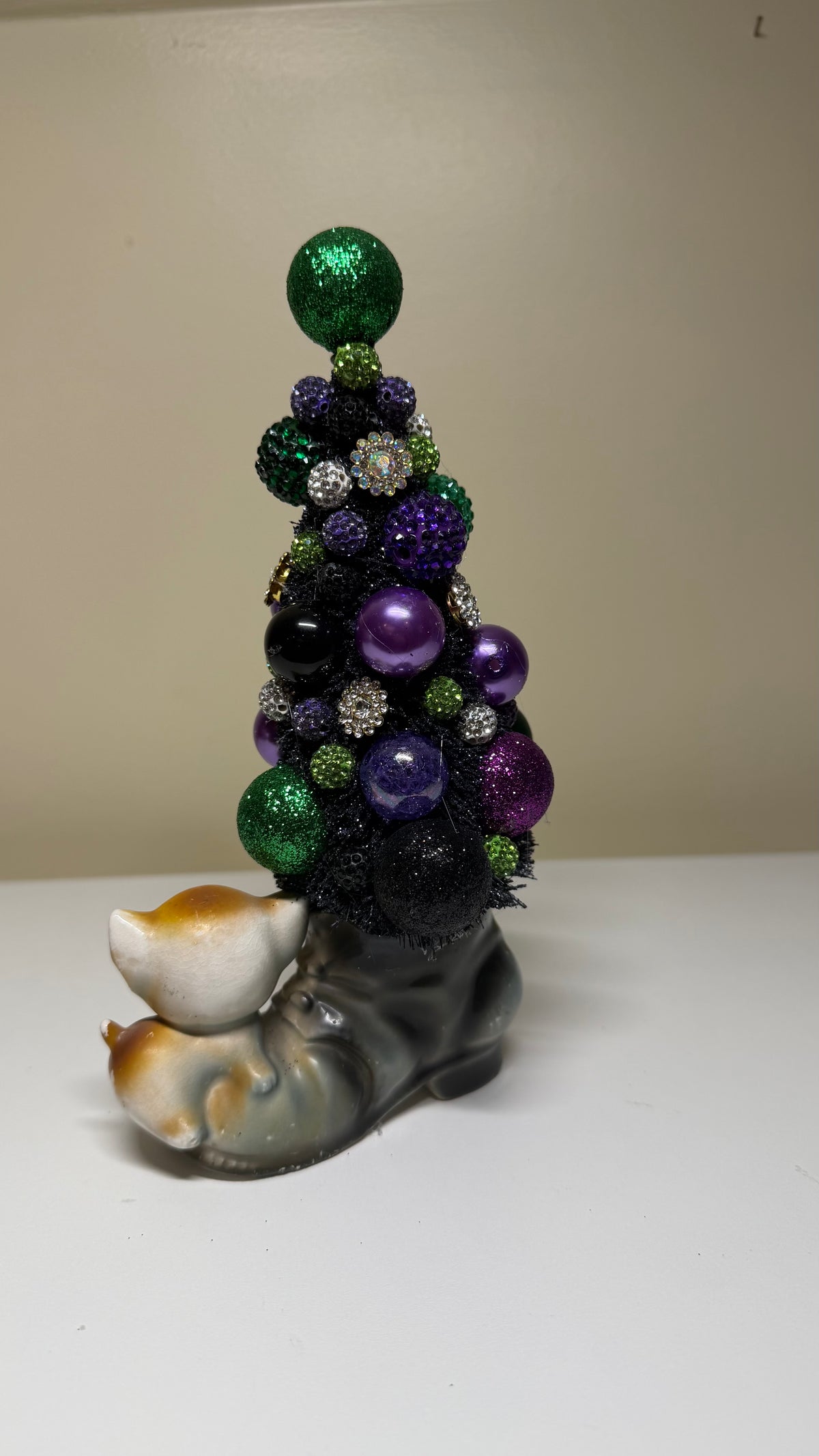 A small decorative Christmas tree sculpture with a gray and white cat figurine at the base, decorated with green, purple, black, and silver ornaments, photographed indoors against a plain beige wall and white surface.