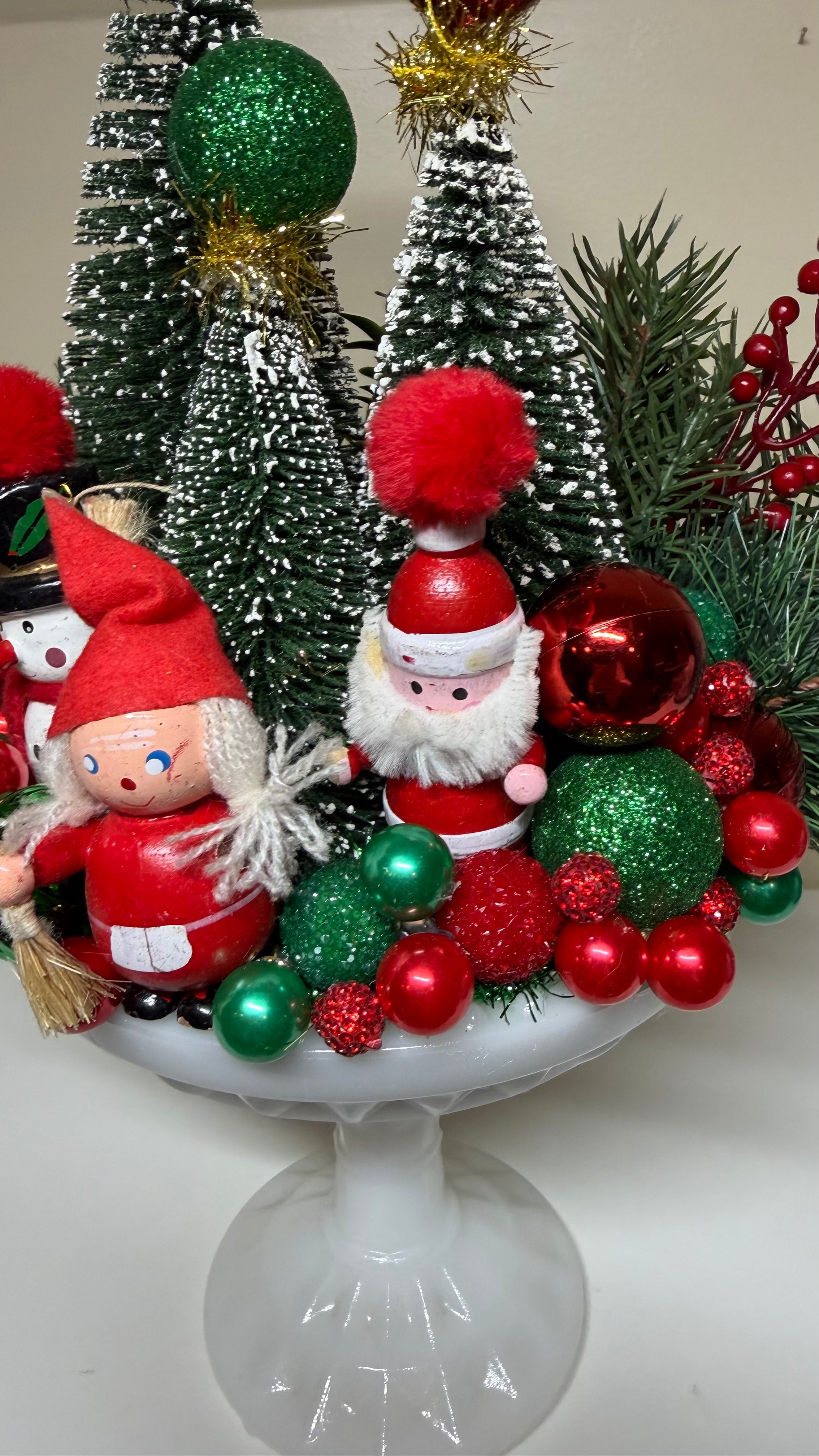 A festive Christmas tabletop decoration arrangement on a white pedestal plate, featuring small artificial Christmas trees, red and green ornaments, a gnome figure, a Santa Claus figure, pine branches, and red berries.