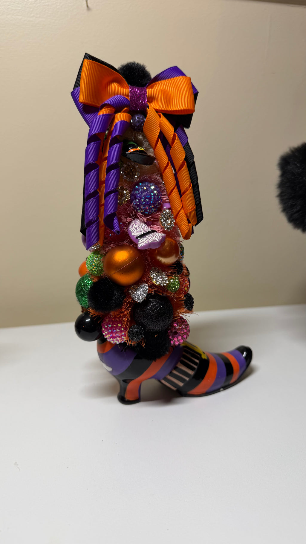 A Halloween-themed boot ornament shaped like a witch's boot, decorated with a large orange and purple striped bow, glittery multicolor embellishments, and striped black, orange, and purple fabric, standing on a white surface against a beige wall.
