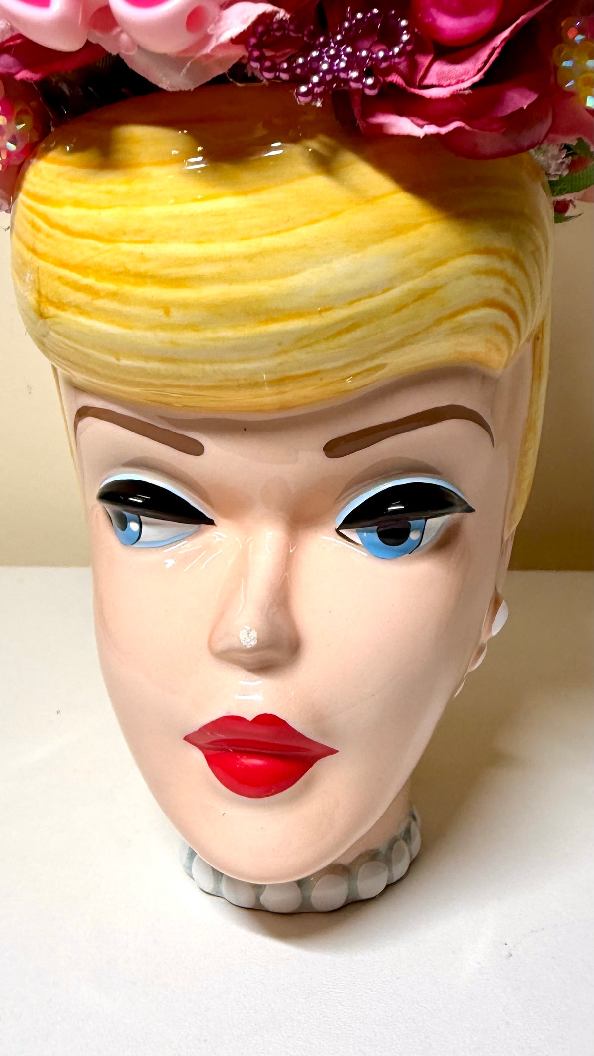 A ceramic decorative head shaped like a woman's face with yellow hair, blue eyes, red lips, and a white pearl necklace, topped with pink and purple flowers.