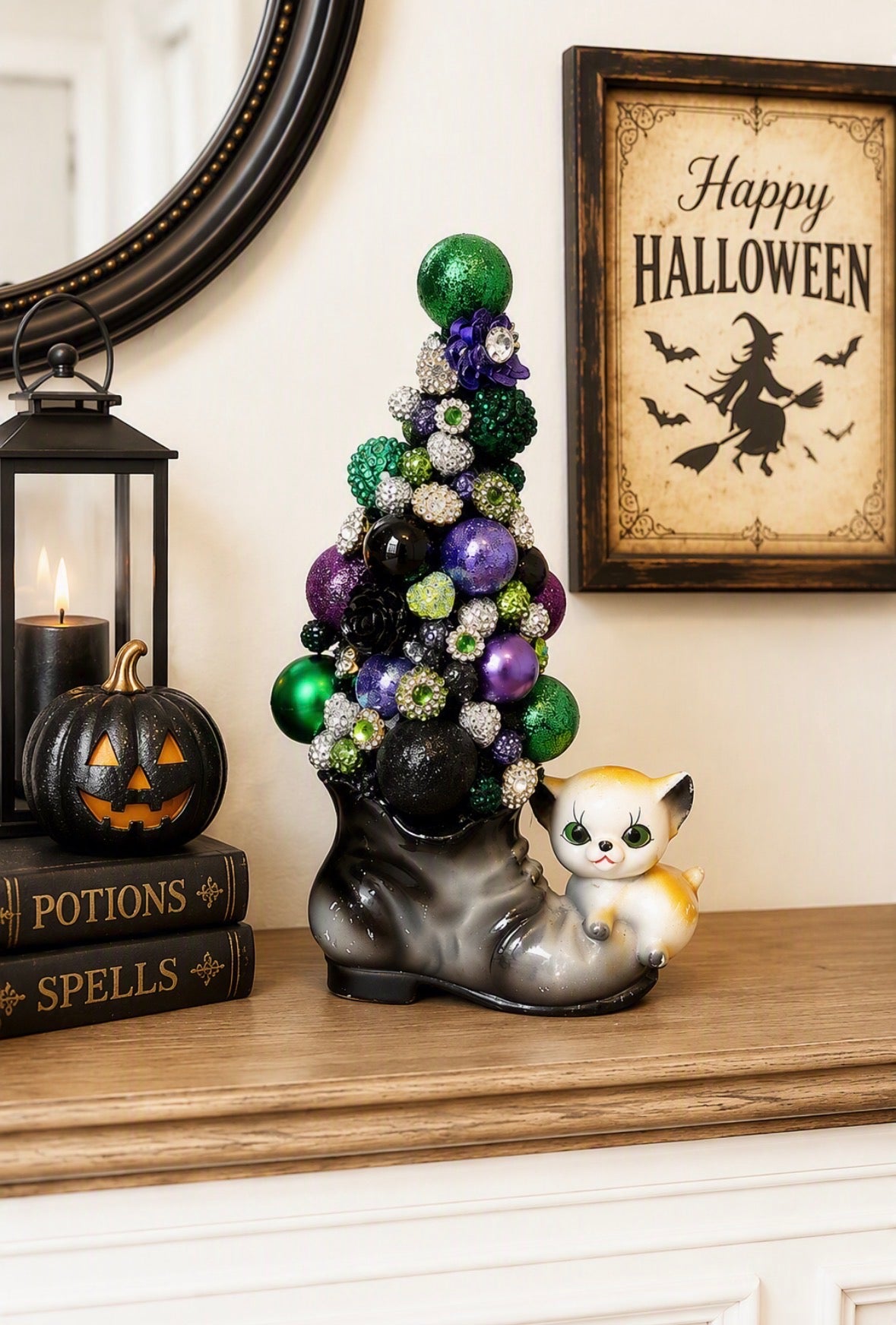 A decorative Halloween display featuring a miniature Christmas tree made of green, purple, black, and silver ornaments, set in a large black boot figurine with a small white and orange cat sitting beside it. The scene is styled on a wooden mantel with a lit lantern, a jack-o'-lantern, books labeled 'Potions' and 'Spells', and a framed 'Happy Halloween' sign on the wall.