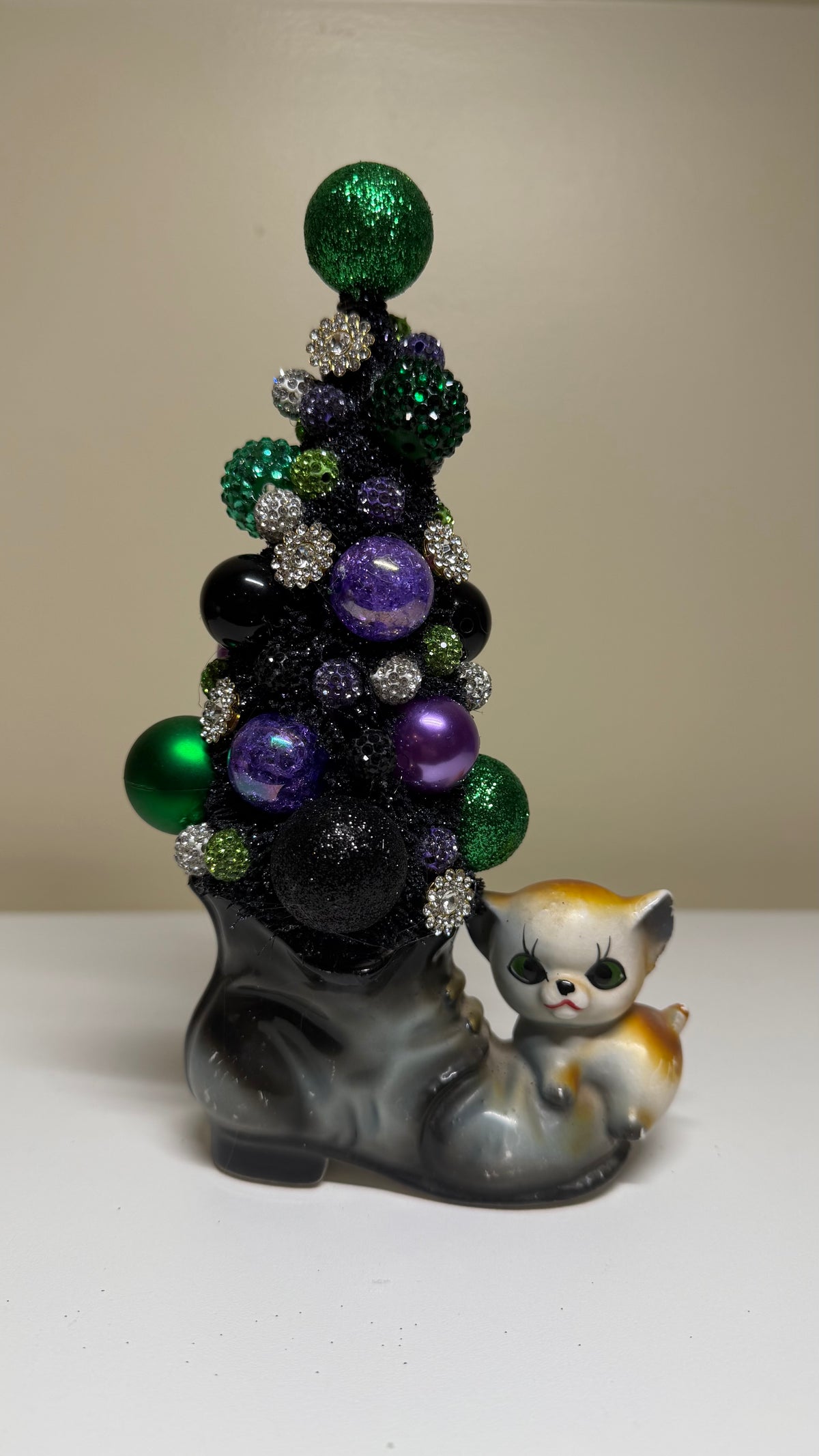 A decorative Christmas tree sculpture made from black glitter and shiny ornaments in green, purple, silver, and black, set in a gray boot-shaped base with a small calico cat figurine beside it, photographed against a plain beige studio background.
