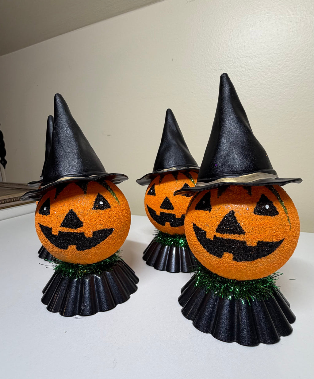 Three Halloween pumpkin decorations shaped like orange jack-o'-lanterns wearing black witch hats, standing on black pleated bases with green tinsel skirts, displayed on a white surface against a beige wall.