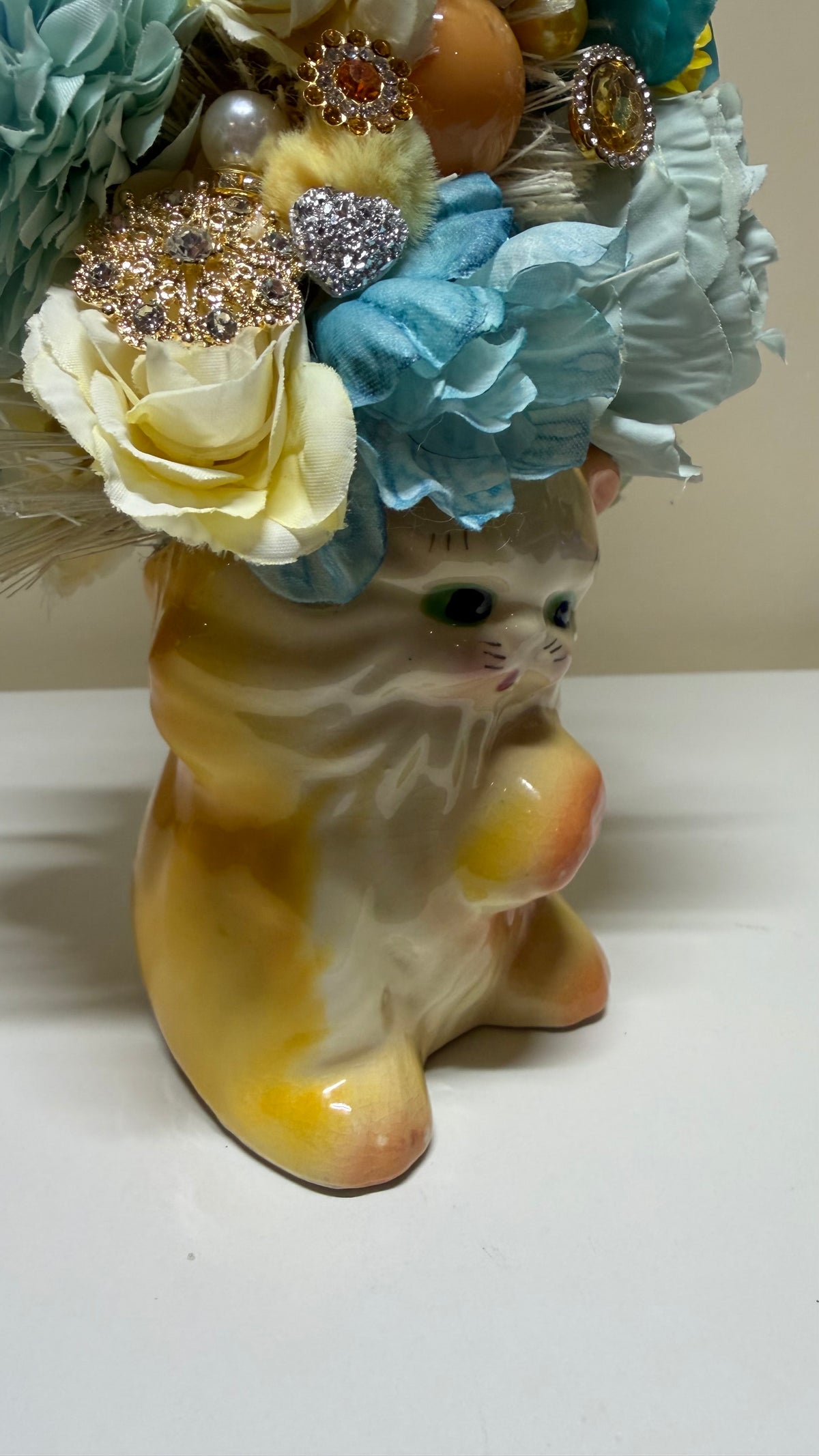 A ceramic cat figurine with a glossy yellow and cream body, decorated with a large floral crown of light blue, cream, and white fabric flowers with jeweled embellishments, sitting on a white surface against a beige background.