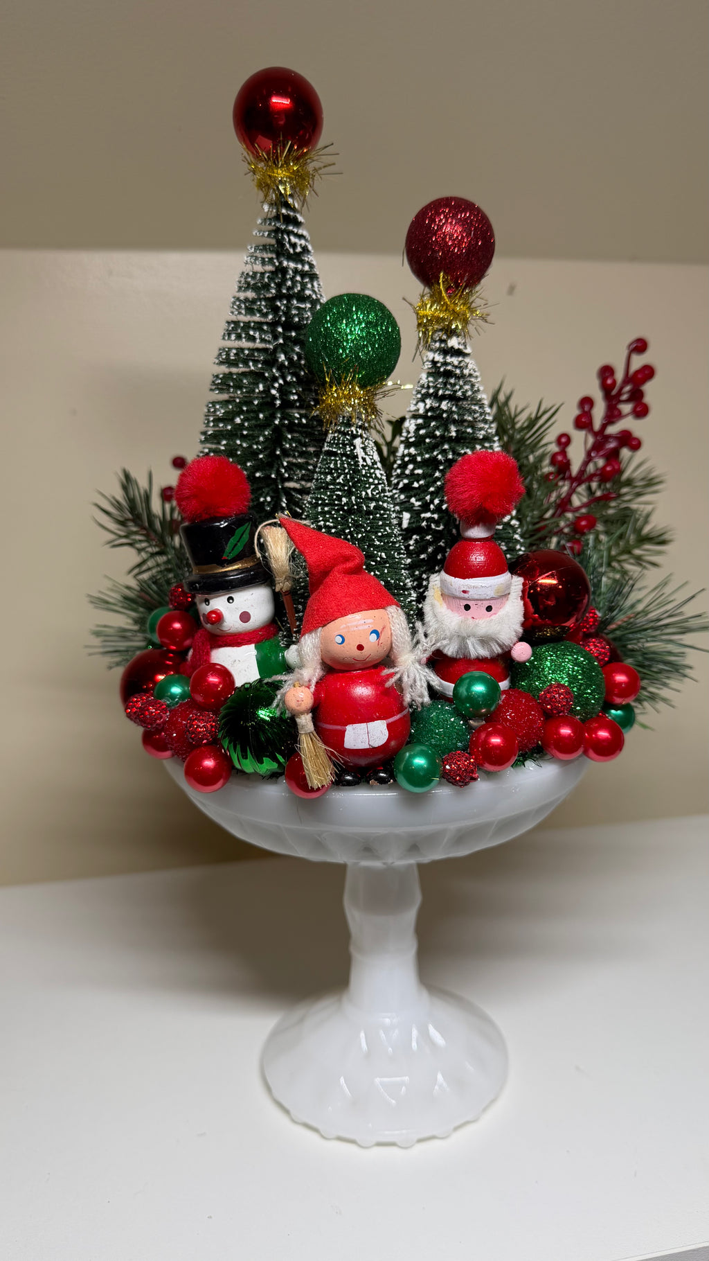 A festive Christmas centerpiece displayed on a white pedestal stand, featuring miniature green Christmas trees, red and green ornaments, pine branches, a snowman, and two Santa Claus figurines.