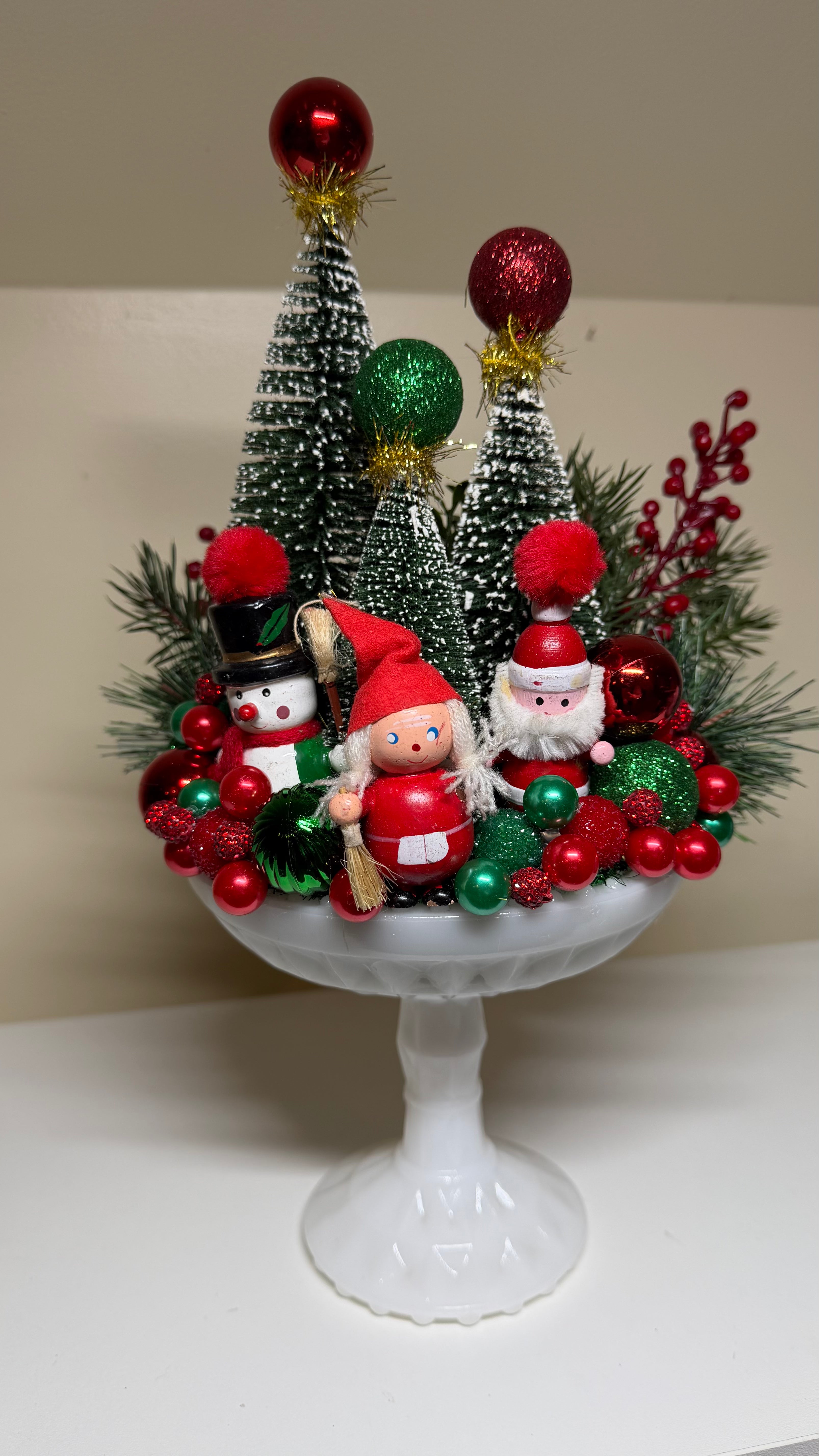 A festive Christmas centerpiece displayed on a white pedestal stand, featuring miniature green Christmas trees, red and green ornaments, pine branches, a snowman, and two Santa Claus figurines.