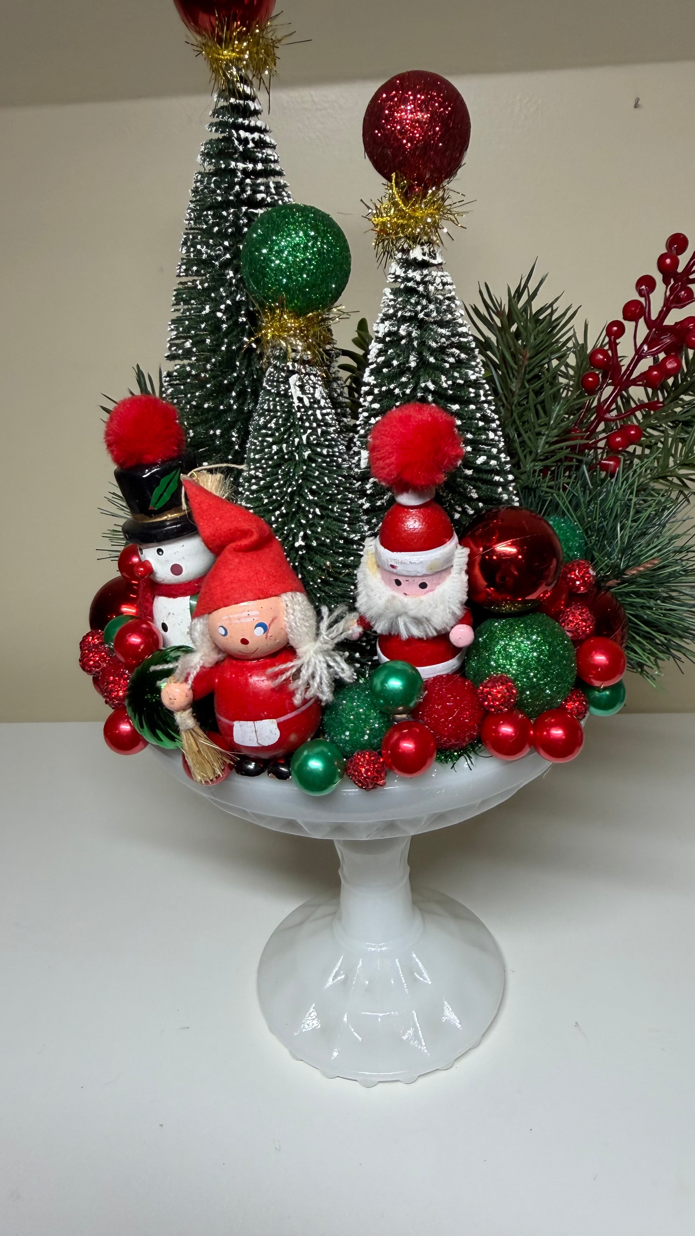 A festive Christmas centerpiece arranged in a white pedestal bowl, featuring miniature silver tree decorations, red and green ornaments, pine branches, and two Santa Claus figurines, photographed against a plain indoor background.