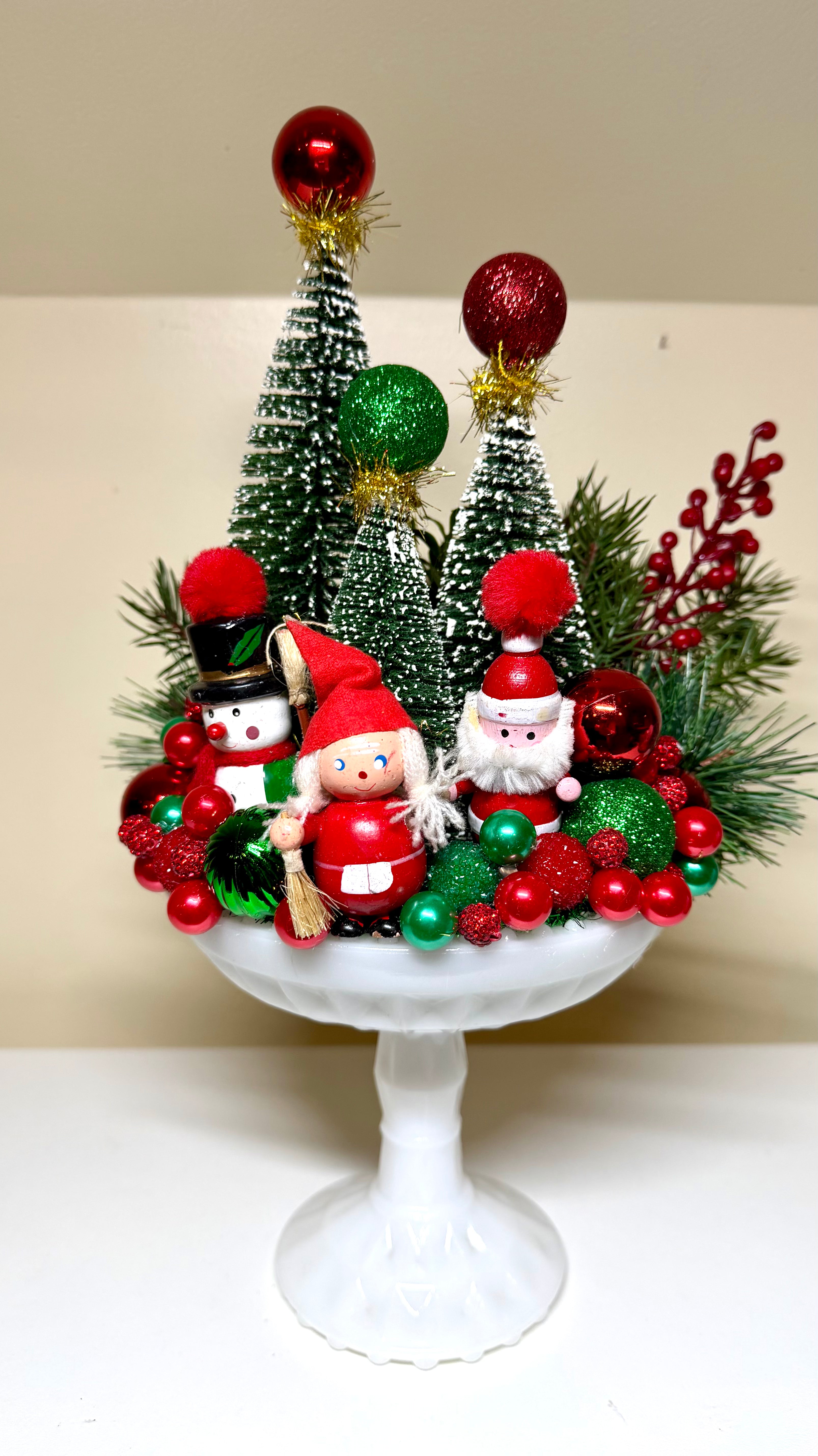 A festive Christmas tabletop centerpiece displayed on a white pedestal stand, featuring miniature green Christmas trees, red and green ornaments, pine branches, berry sprigs, and two small Santa-themed figurines.