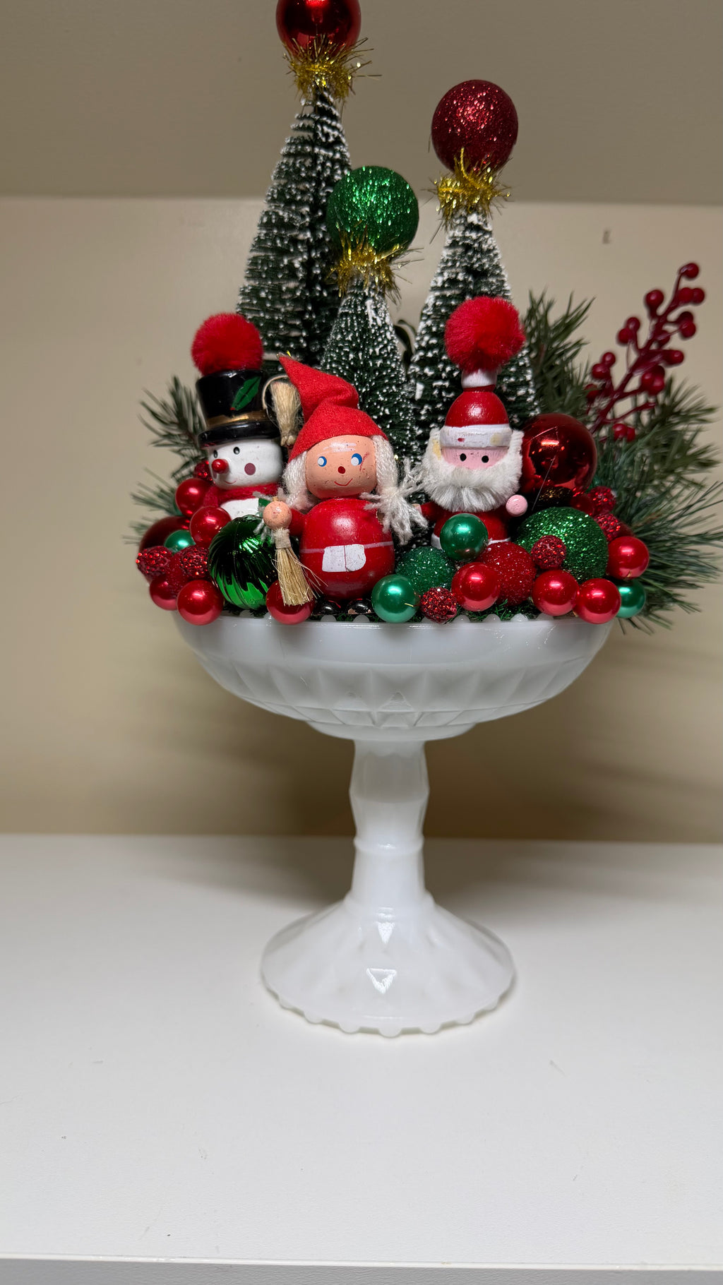 A festive Christmas tabletop centerpiece in a white pedestal bowl, decorated with miniature green Christmas trees, red and green ornaments, pine branches, and small Santa Claus and snowman figurines, set against a plain beige wall and white surface.