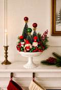 A festive Christmas tabletop centerpiece in a white pedestal bowl, featuring miniature green trees, red and green ornaments, pine branches, berries, and small Santa-themed figurines, styled on a mantel with a candle, stockings, and framed wall art.