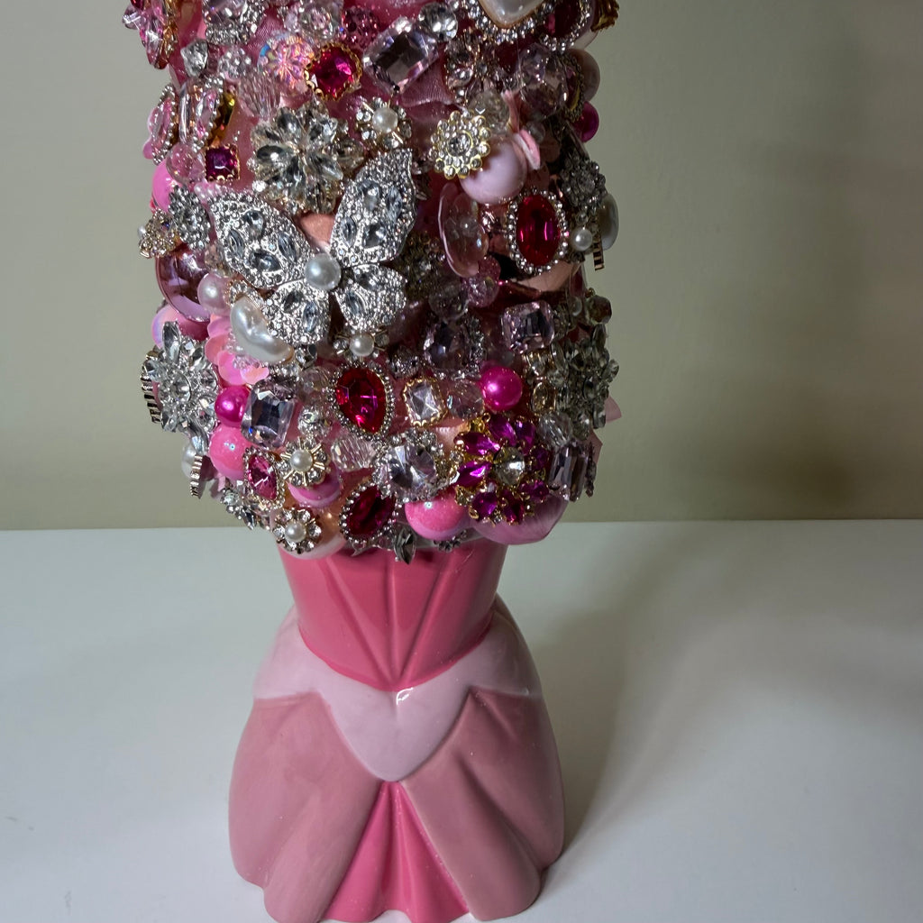 A pink ceramic vase decorated with an elaborate arrangement of pink, silver, and clear rhinestones, brooches, and faux pearls, set against a plain beige background.