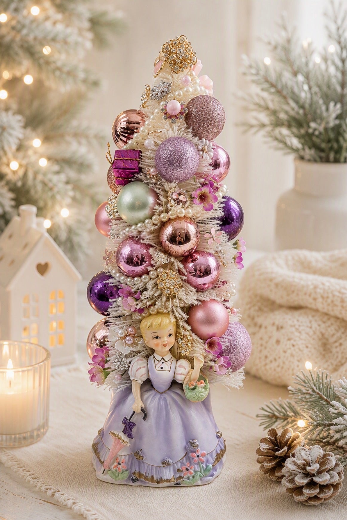 A decorative miniature Christmas tree with a blonde doll dressed in a lavender gown at the base, surrounded by pastel pink, purple, and green ornaments on a soft cream surface with holiday lights and plants in the background.
