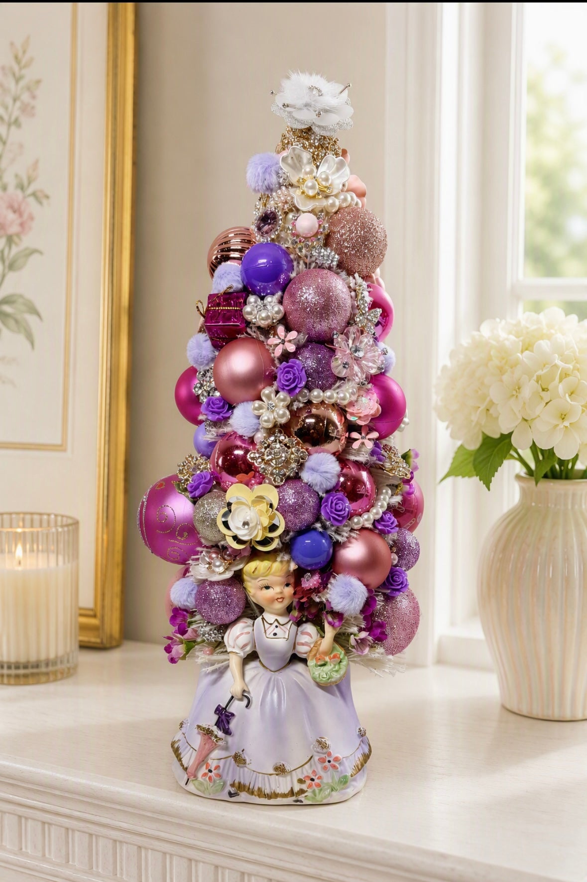 A decorative Christmas tree ornament featuring a miniature tree adorned with pink, purple, and gold ornaments, flowers, and a princess figurine at the base, displayed on a windowsill with a candle and flowers nearby.