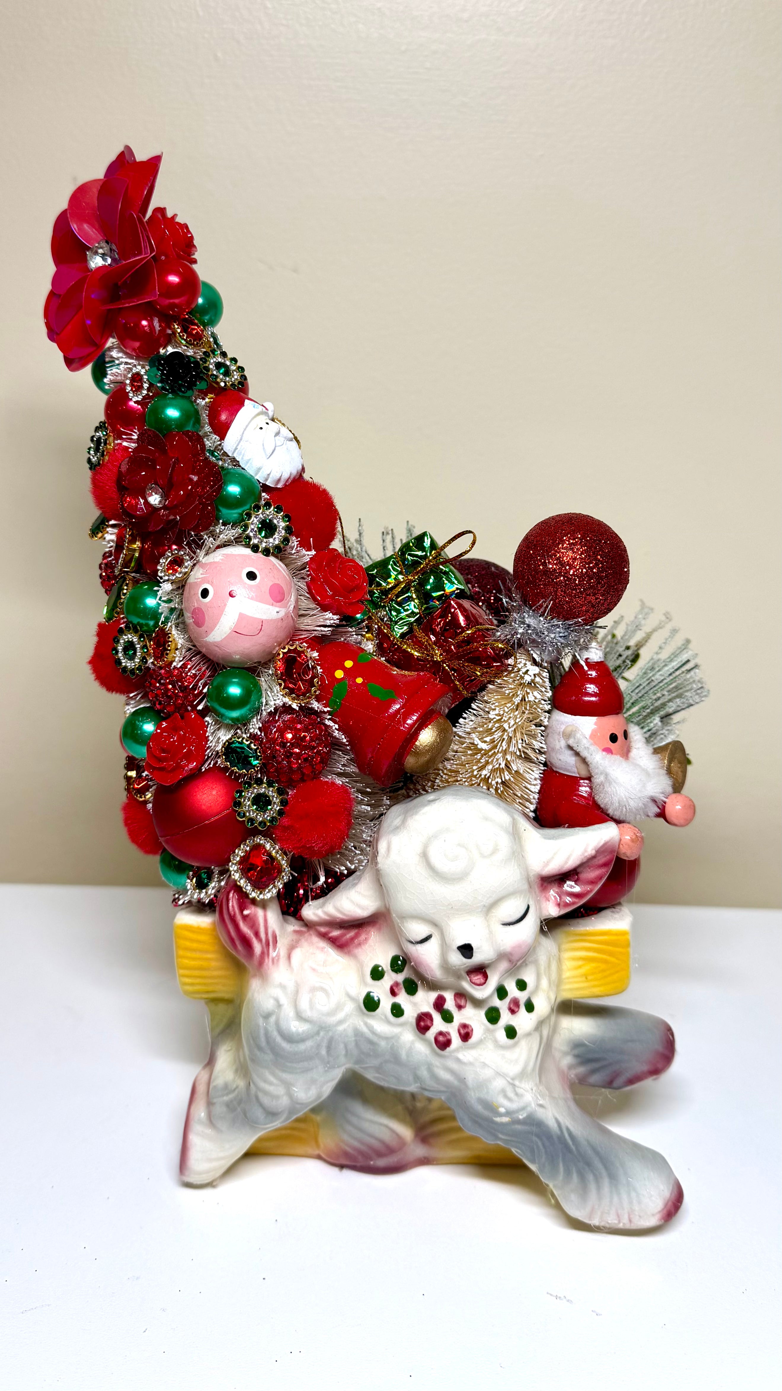 A festive Christmas ornament display featuring a white ceramic lamb figurine carrying a tall, dense Christmas tree decorated with red and green ornaments, red baubles, a small gold tree, and Santa Claus figures, set against a plain beige wall and white surface.