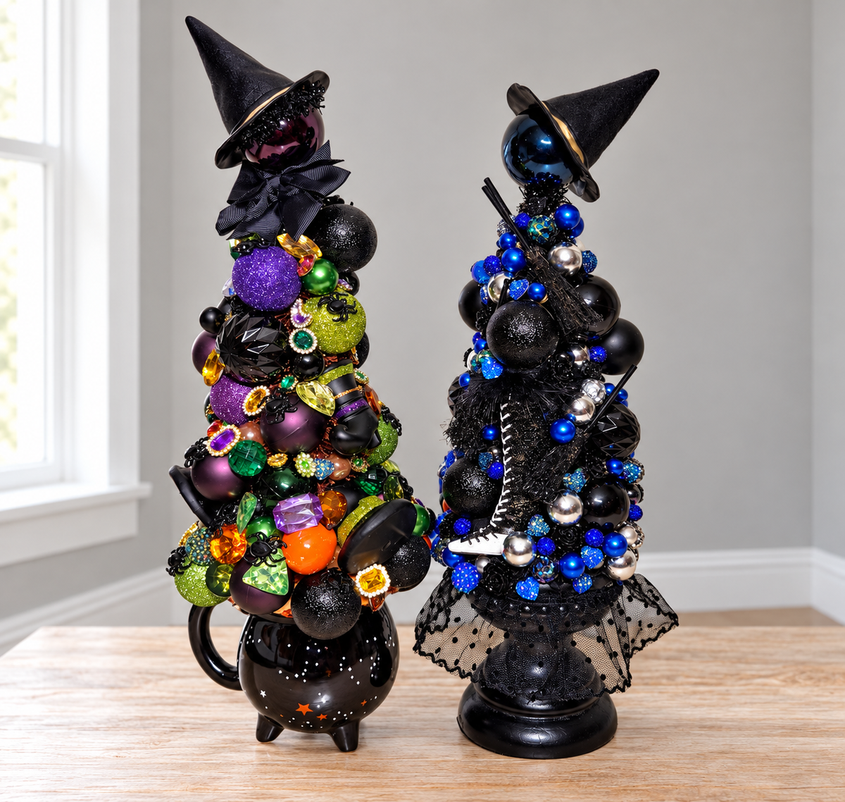 Two decorative black witch-themed Christmas trees on a light wood tabletop in front of a window. The left tree is shaped like a cauldron and is decorated with black ornaments, purple and green glittery baubles, and small colorful accents, topped with a black witch hat and black bow.